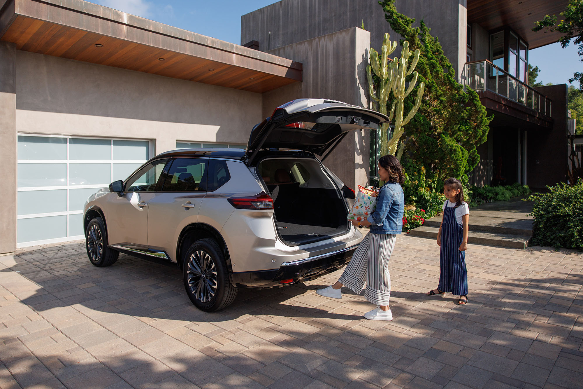 An adult puts a gift into the open cargo space of a tan Nissan Rogue with a child watching.