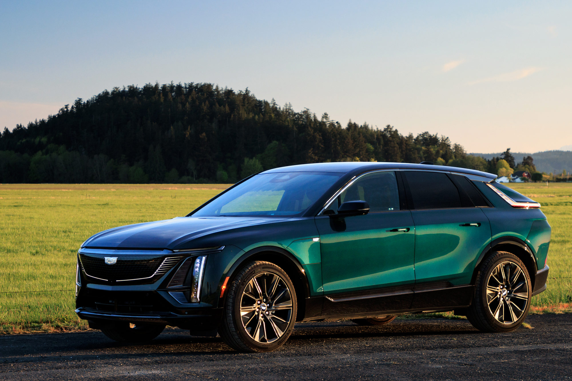 Front-quarter of a blue-green 2024 Cadillac Lyriq parked by a field with a tree-covered hilltop