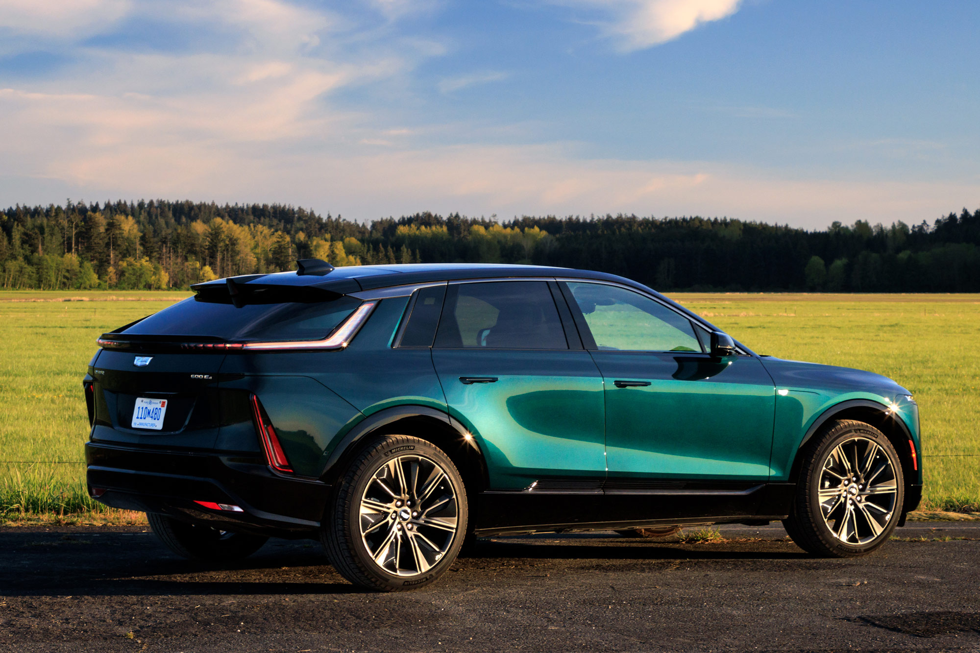 Rear-quarter of a blue-green 2024 Cadillac Lyriq parked by a field with a tree-covered hilltop