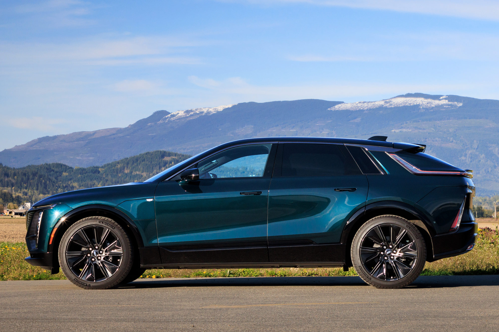 Side view of a blue-green 2024 Cadillac Lyriq parked on the pavement with a field and mountains in the background