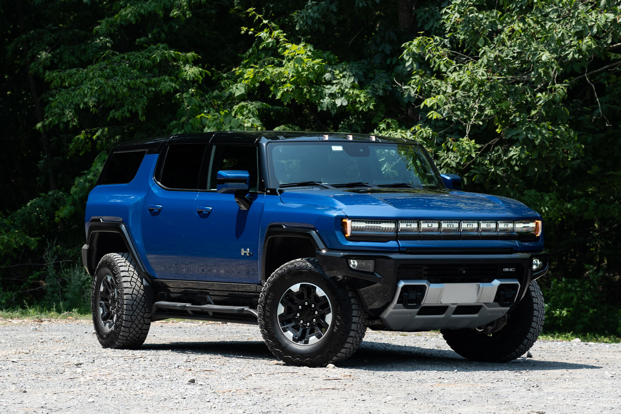 Front-quarter of a Supernova blue 2024 GMC Hummer EV SUV 3X parked on gravel with a forest in the background