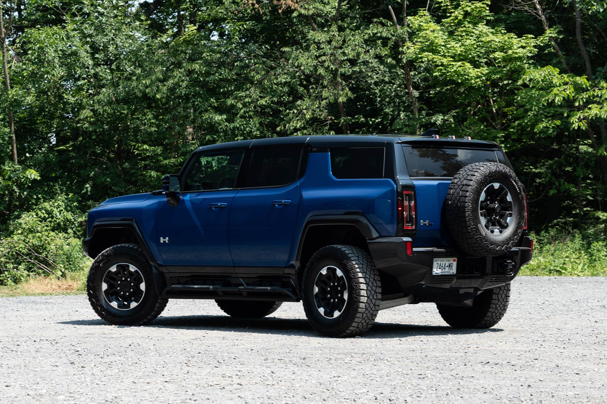 Rear-quarter of a Supernova blue 2024 GMC Hummer EV SUV 3X parked on gravel with a forest in the background