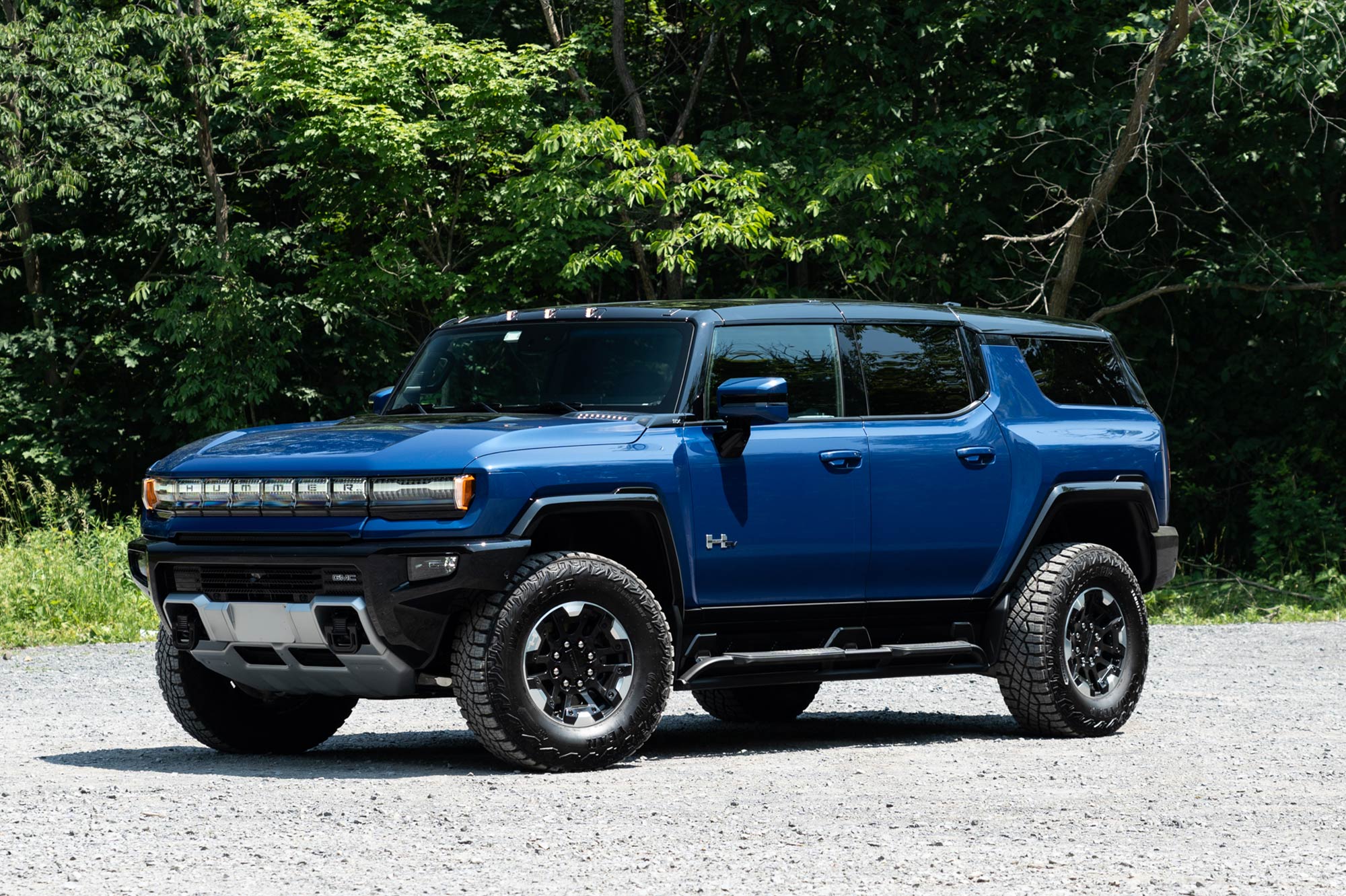 Front-quarter of a Supernova blue 2024 GMC Hummer EV SUV 3X parked on gravel with a forest in the background