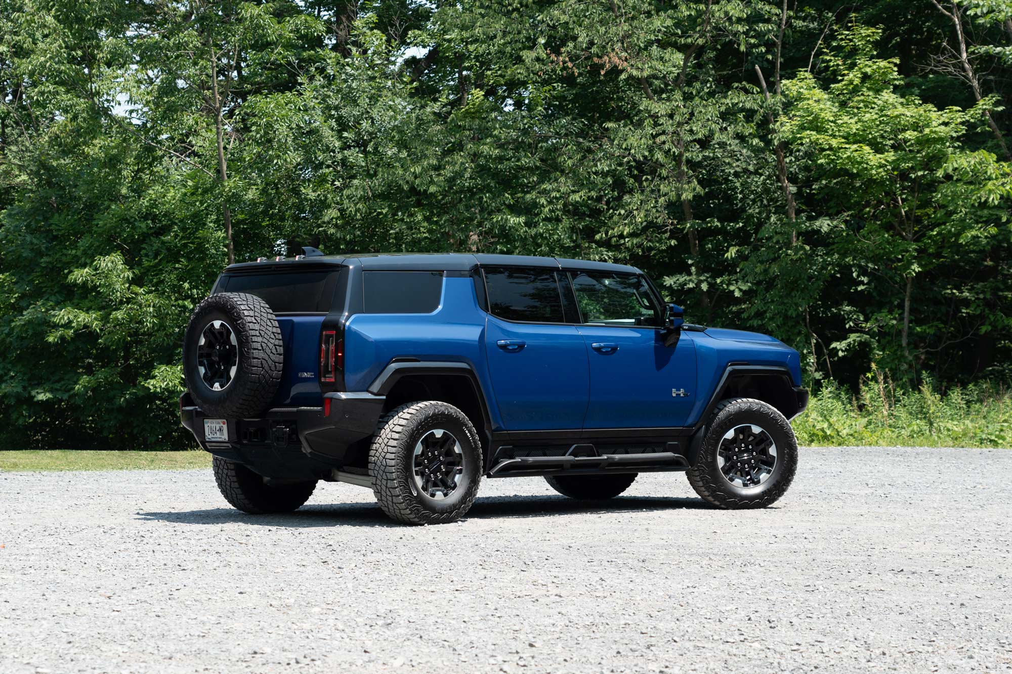 Rear-quarter of a Supernova blue 2024 GMC Hummer EV SUV 3X parked on gravel with a forest in the background