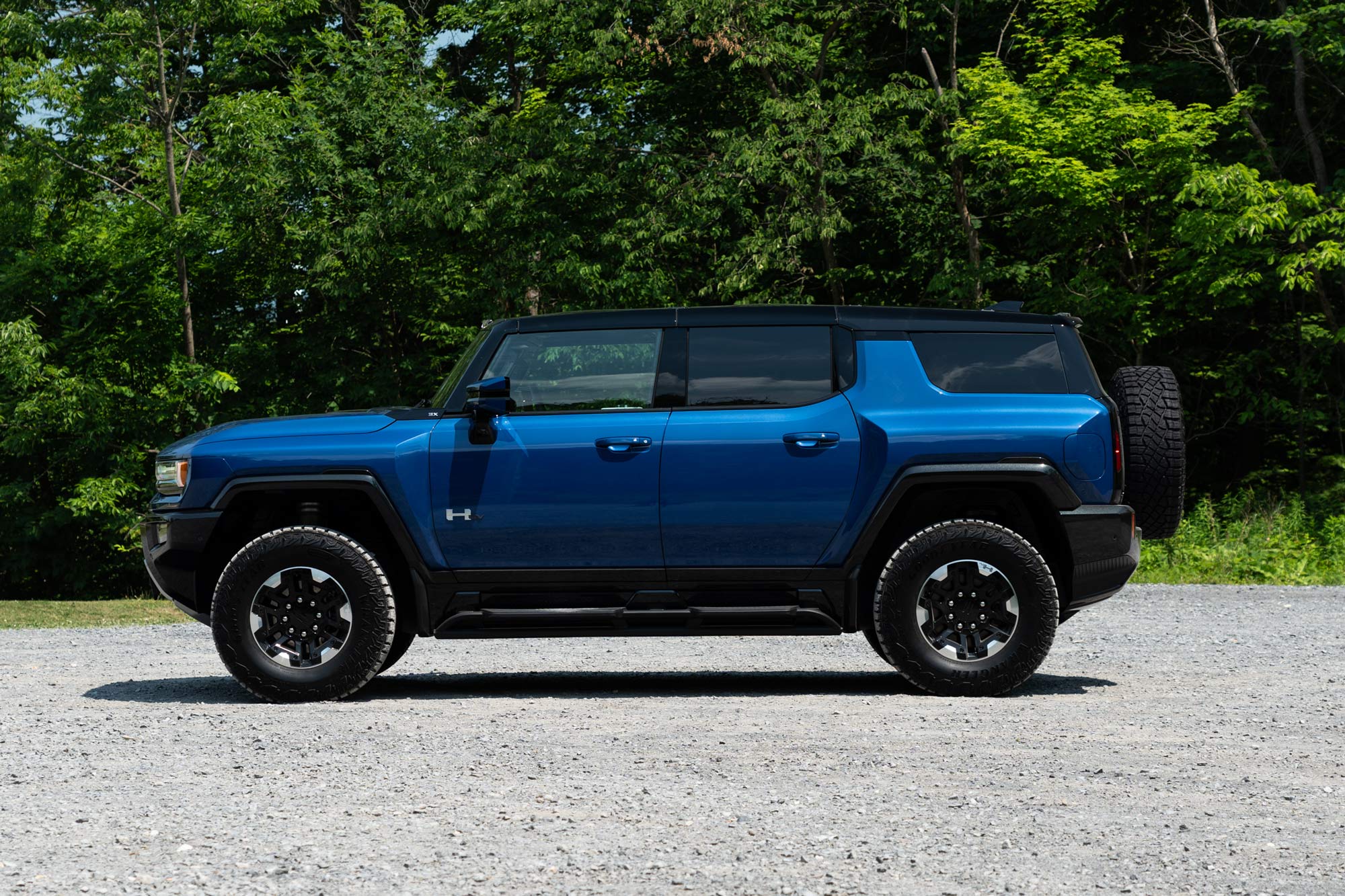 Side view of a Supernova blue 2024 GMC Hummer EV SUV 3X parked on gravel with a forest in the background