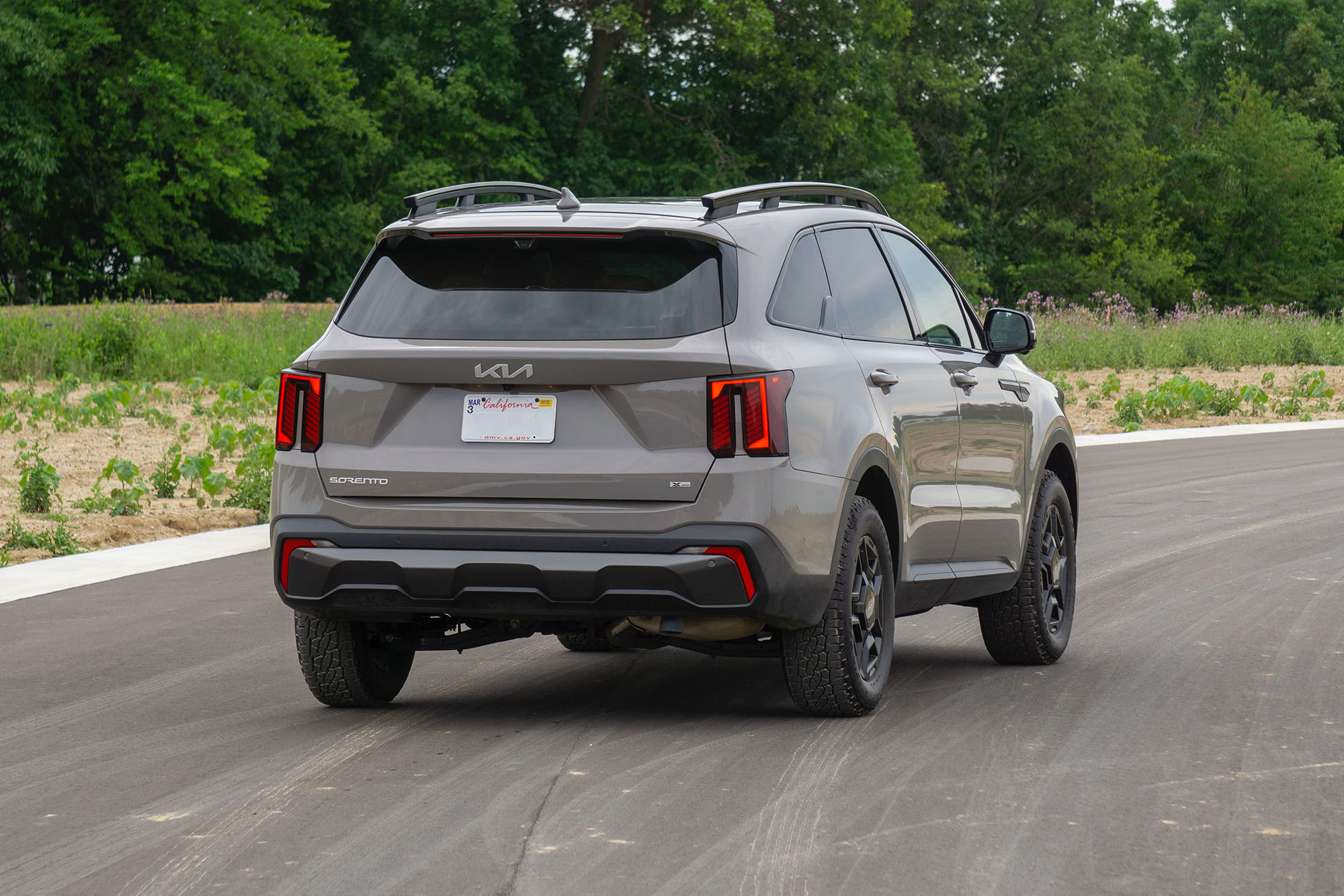 Rear-quarter view of a Road Rider Brown 2024 Kia Sorento SX Prestige X-Pro parked on a road with trees in the background.