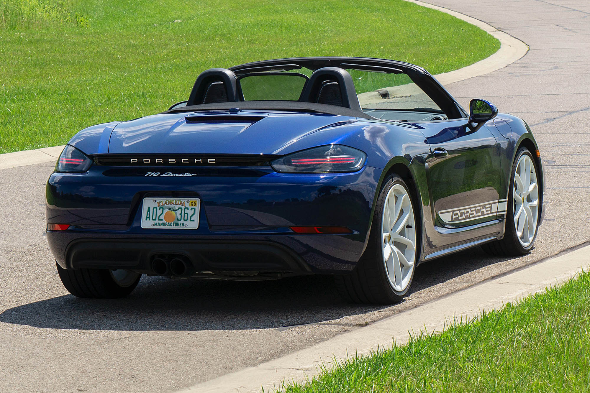 Rear-quarter view of a blue 2024 Porsche 718 Boxster Style parked on the pavement in front of an S-curve