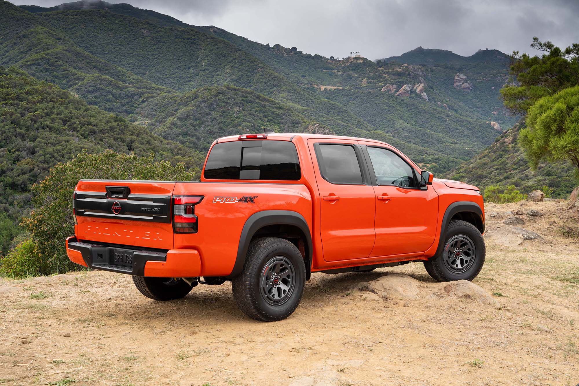 Right-rear view of an orange 2025 Nissan Frontier Pro-4X parked on dirt in front of mountains 