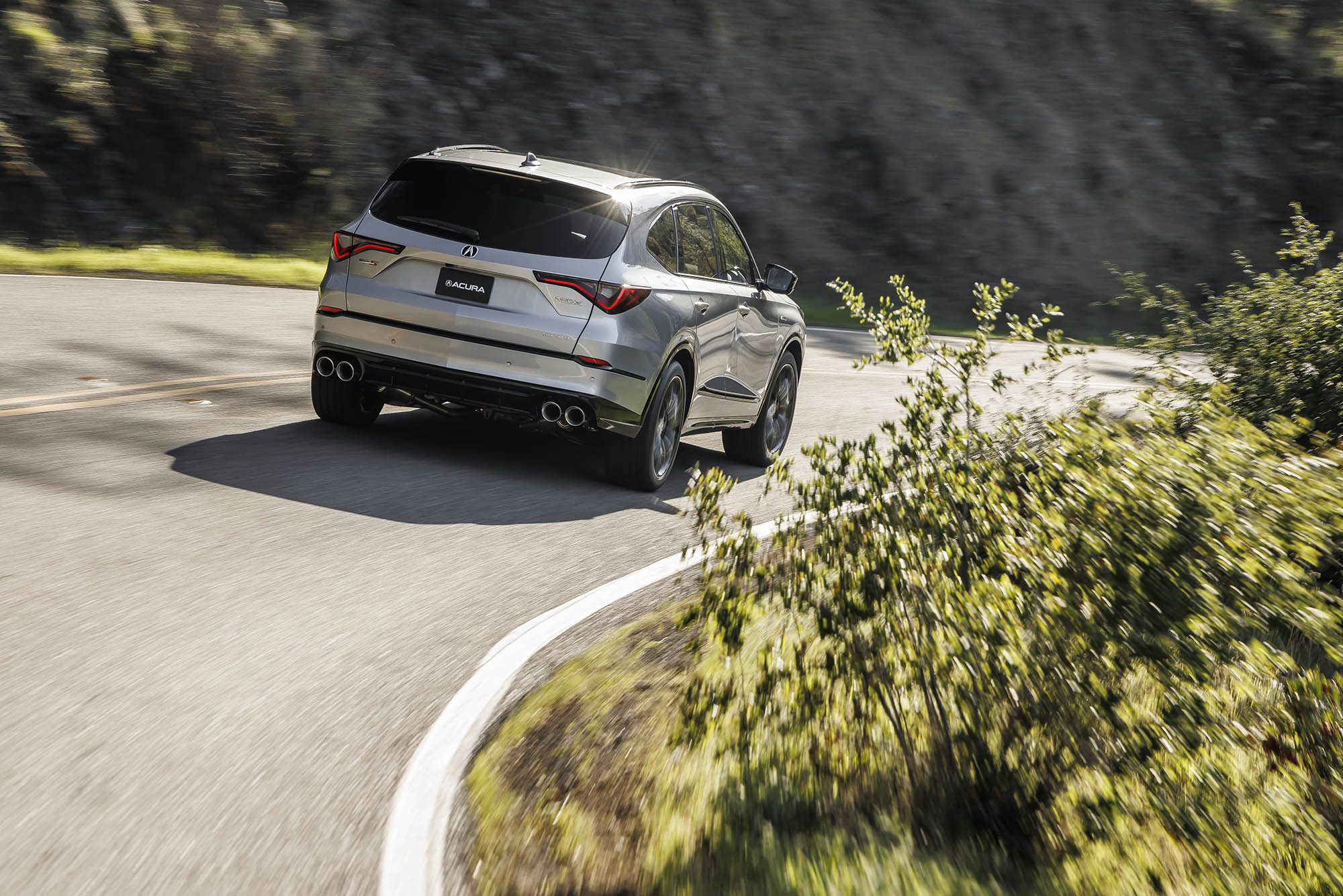 Rear view of Acura MDX in silver driving on a curving paved road.