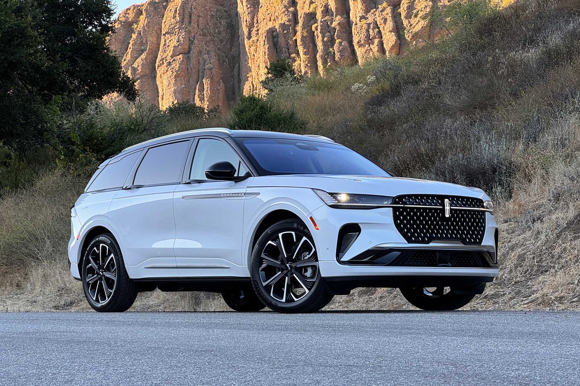 Front-quarter view of a white 2024 Lincoln Nautilus Reserve parked on a road with a brush-covered hill and a sunlit bluff in the background.