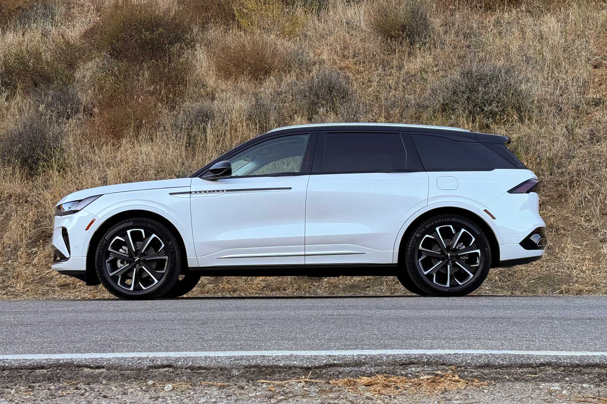 Side view of a white 2024 Lincoln Nautilus Reserve parked on a road with a brush-covered hill in the background.