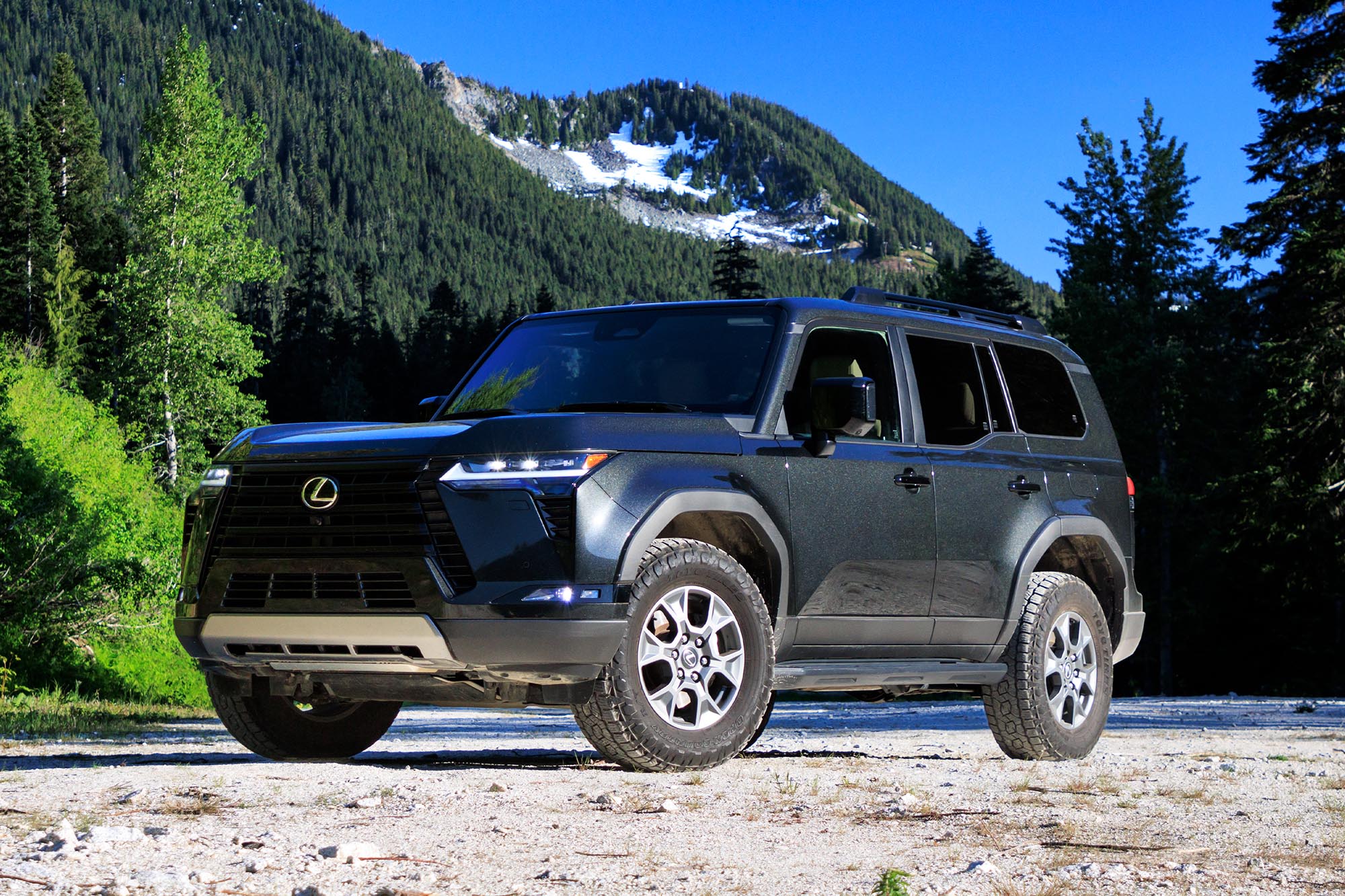 Front-quarter view of a dark gray Caviar 2024 Lexus GX Overtrail parked on a trail with a snow-capped mountain in the background.