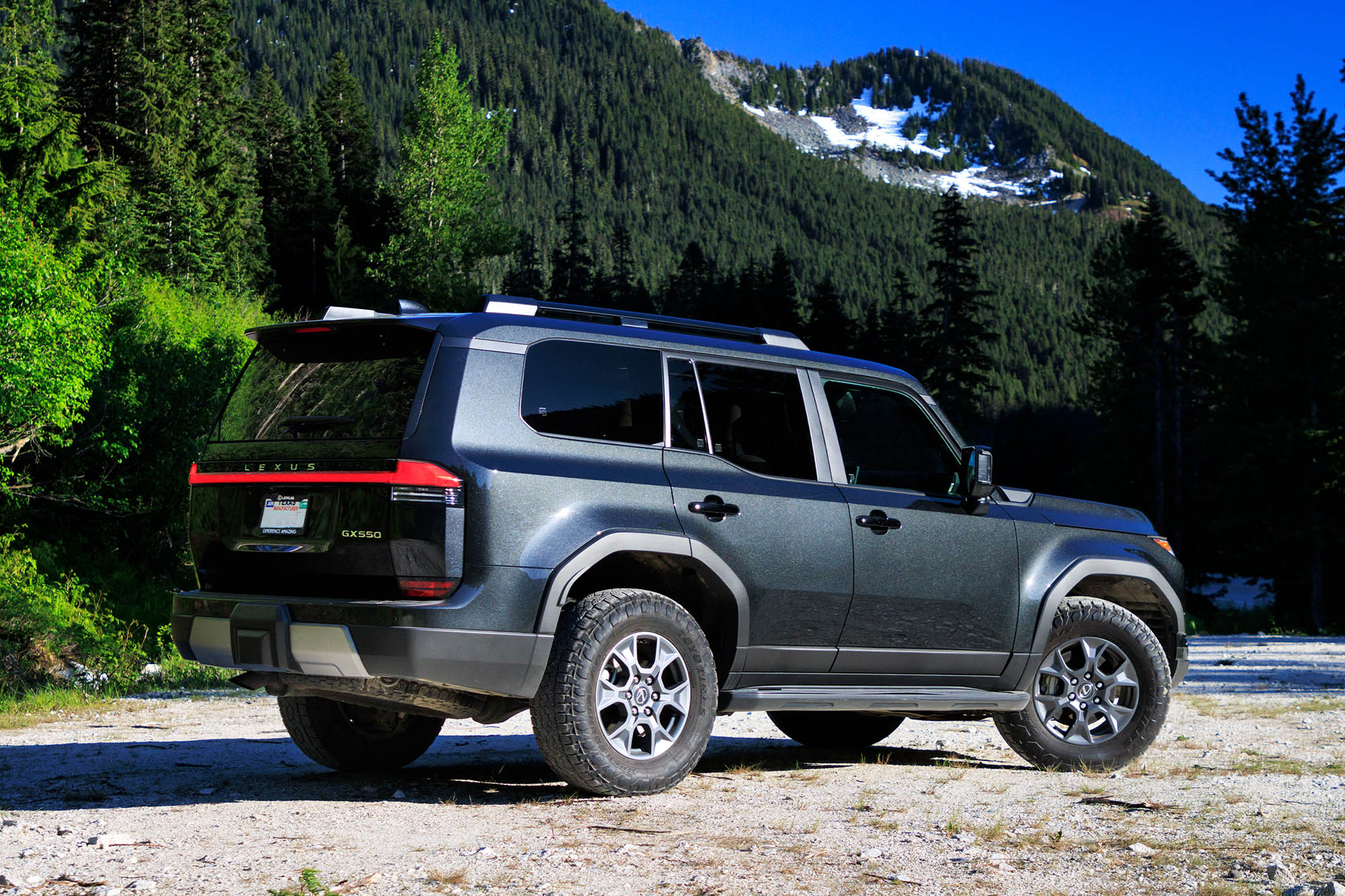 Rear-quarter view of a dark gray Caviar 2024 Lexus GX Overtrail parked on a trail with a snow-capped mountain in the background.