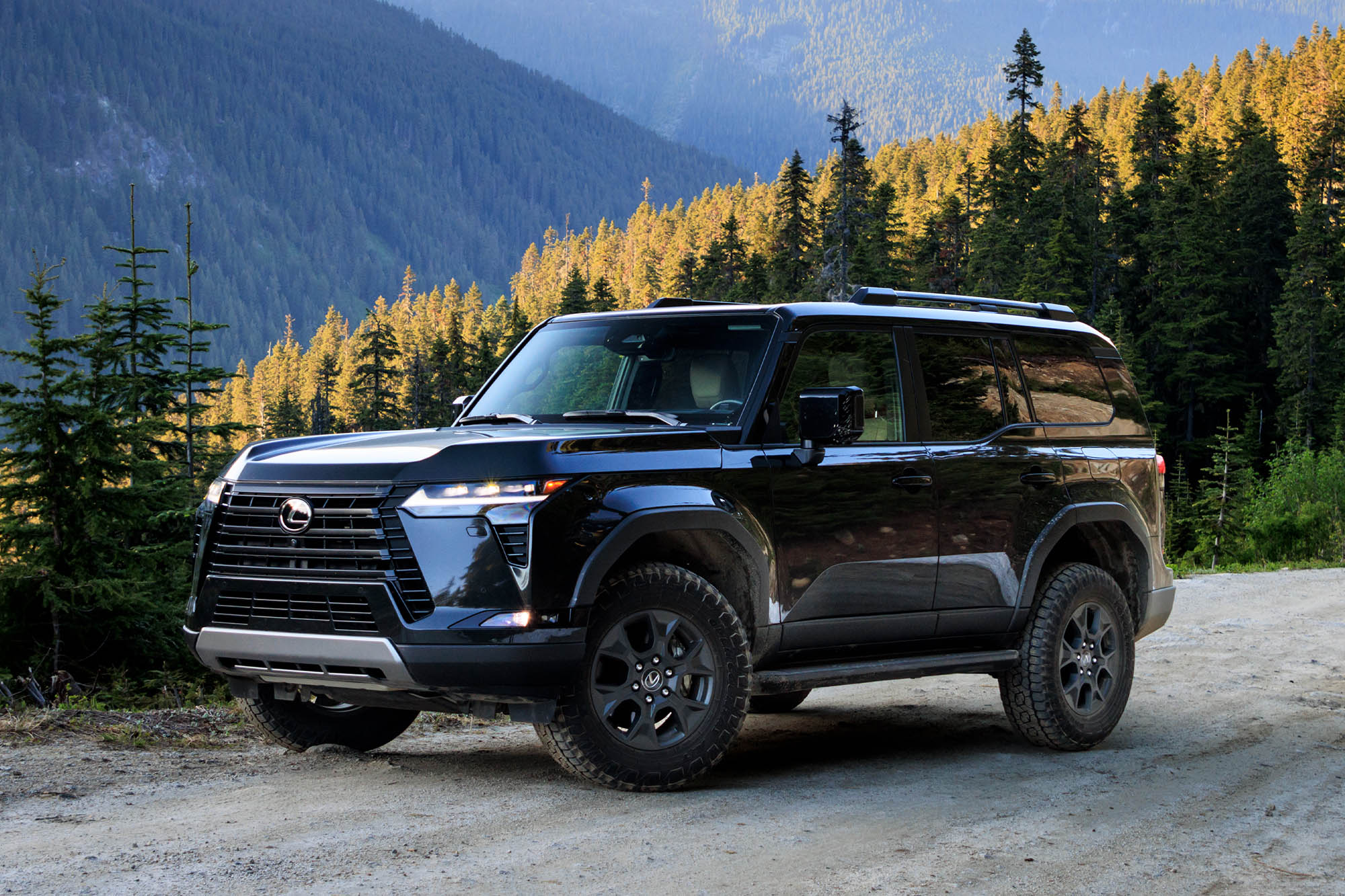 Front-quarter view of a dark gray Caviar 2024 Lexus GX Overtrail parked on a trail with pine tree-covered mountains in the background.