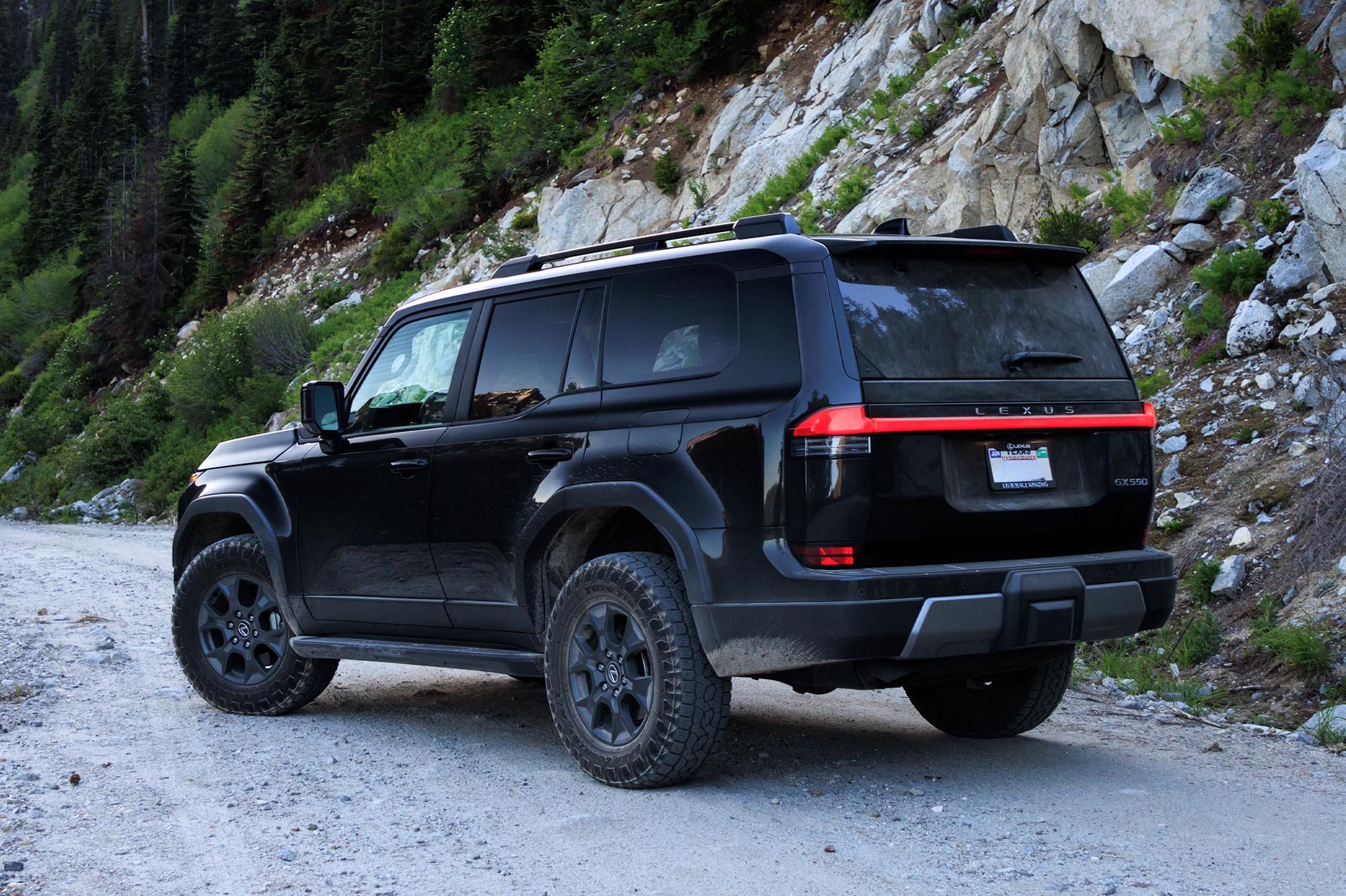 Rear-quarter view of a dark gray Caviar 2024 Lexus GX Overtrail parked on a trail with a rock and tree-covered hillside in the background.