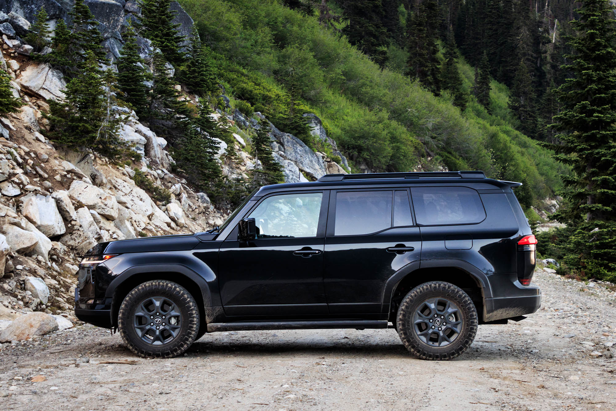 Side view of a dark gray Caviar 2024 Lexus GX Overtrail parked on a trail with a rock and tree-covered hillside in the background.