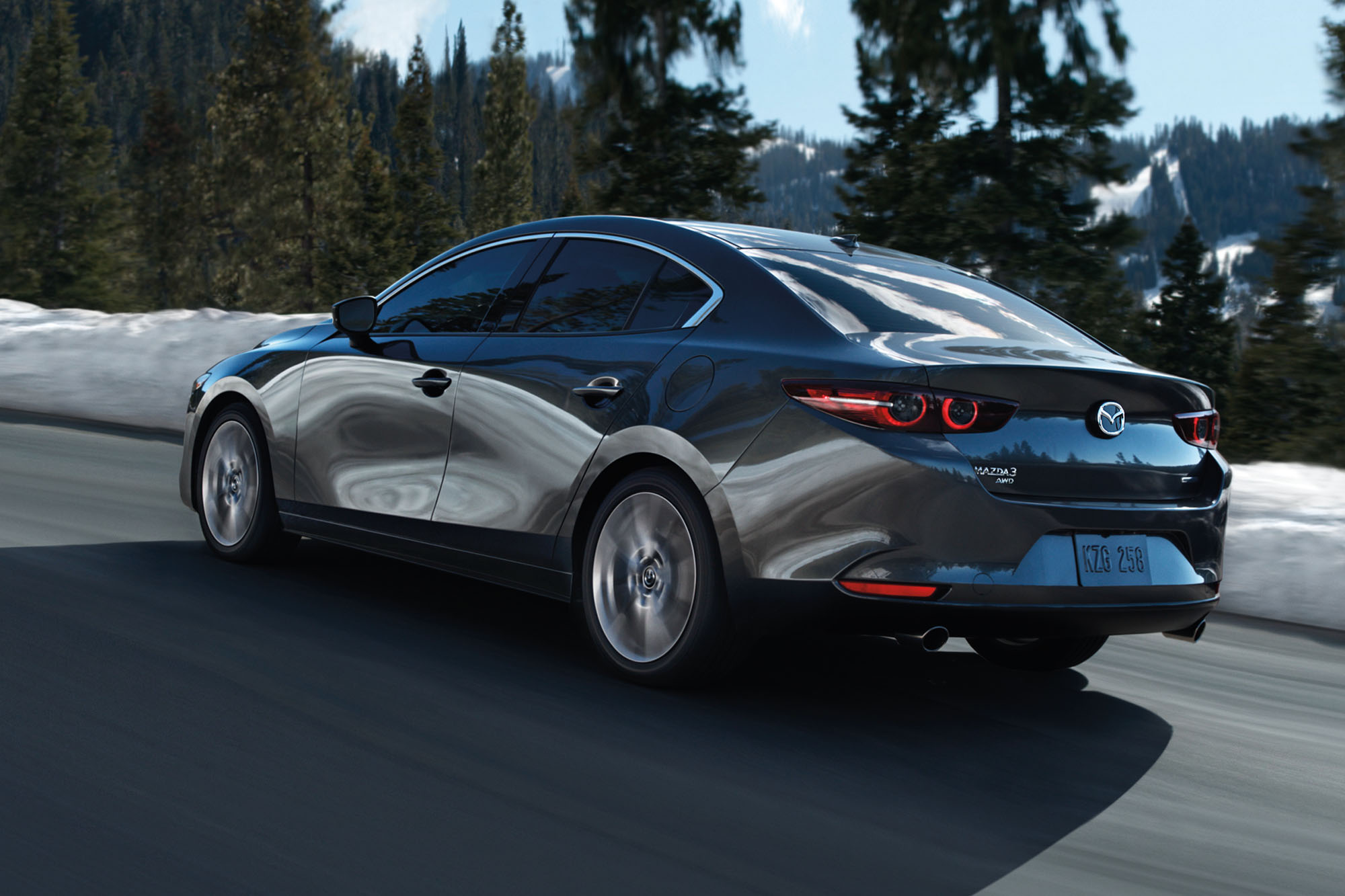 Rear three-quarter view of a dark silver Mazda 3 driving on a paved road with a snow bank on the side.