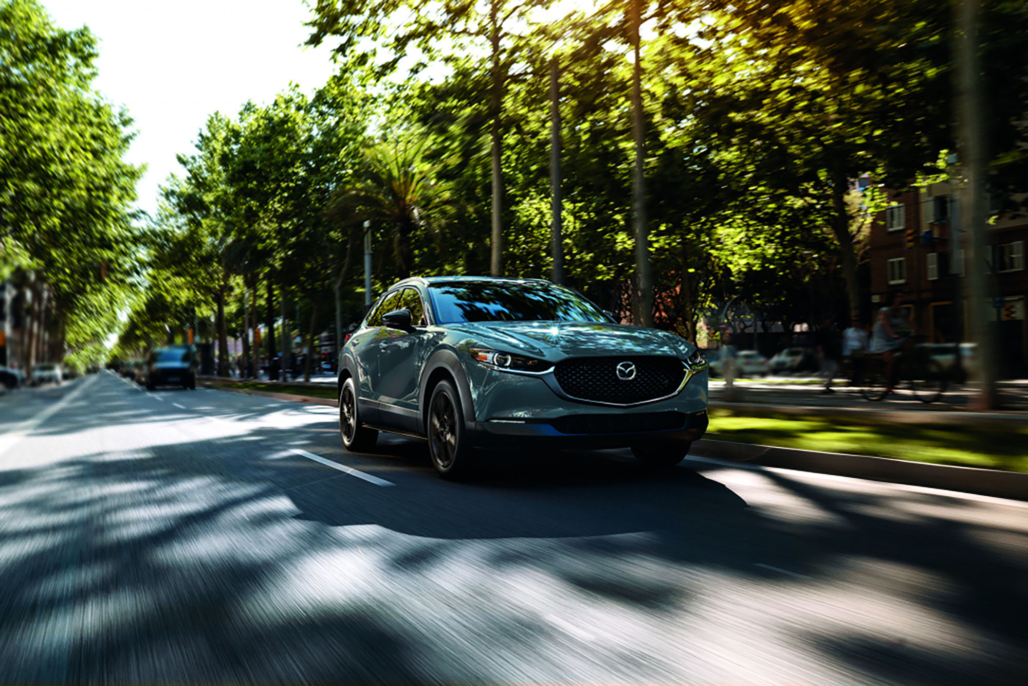 Blue-gray Mazda CX-30 driving on a paved road past trees.