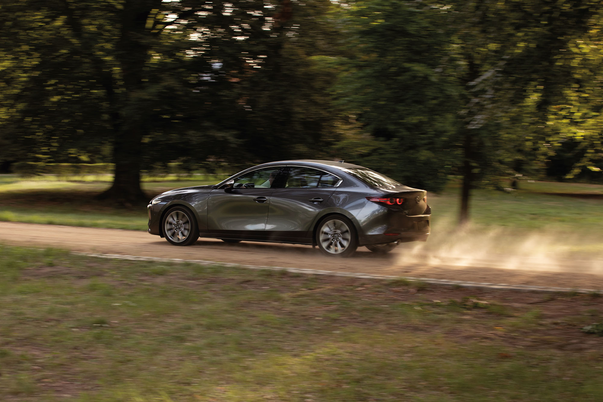 Dark silver Mazda 3 sedan driving on a dirt road past trees.