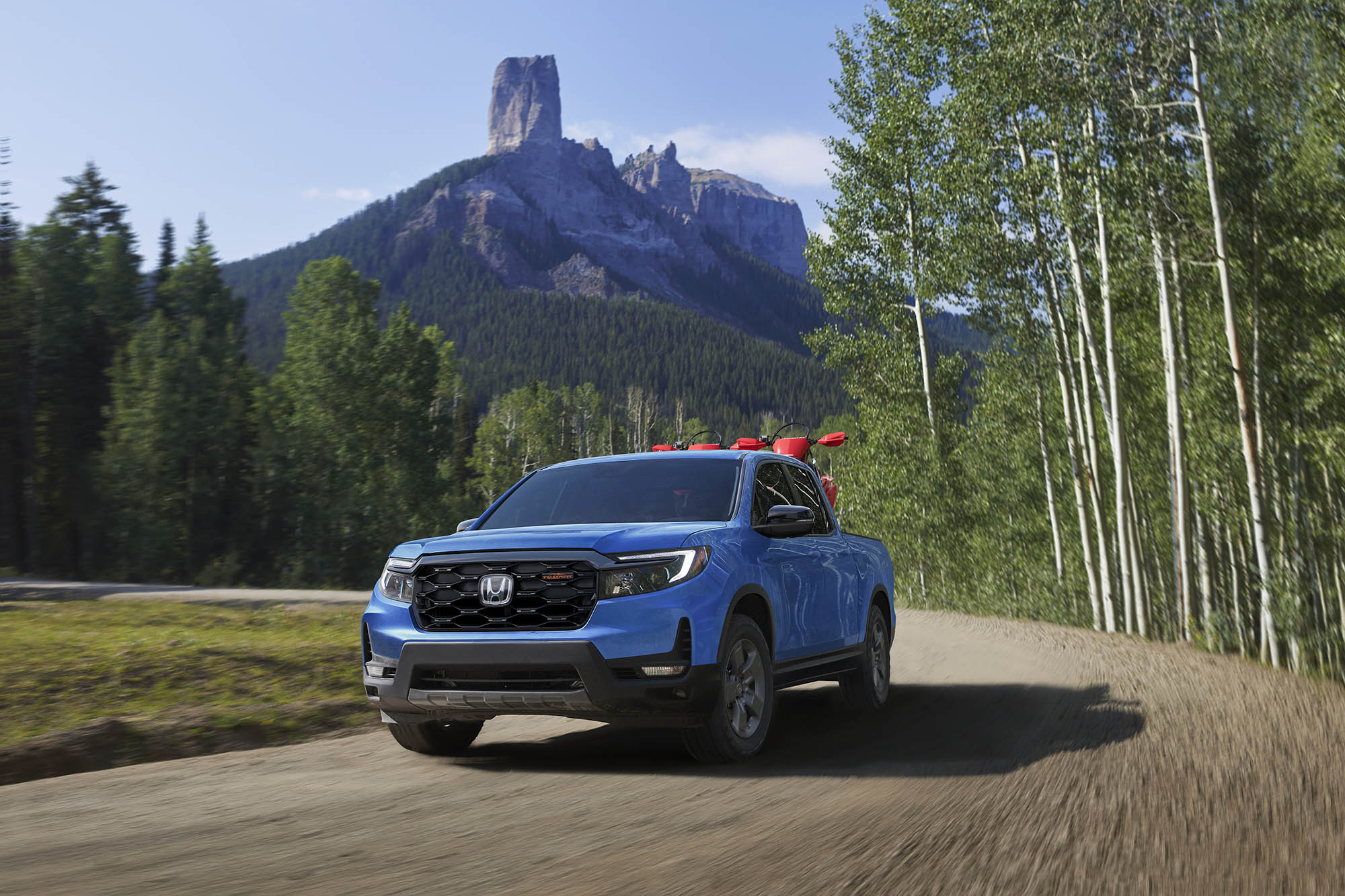 Blue Honda Ridgeline driving on a dirt road with mountains behind.