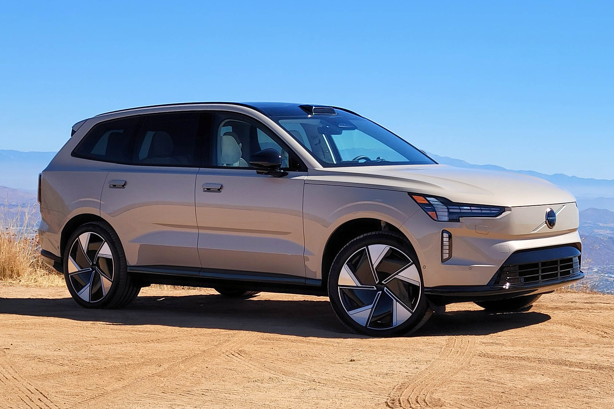 2025 Volvo EX90 Ultra in Sand Dune parked on dirt with a distant valley and blue sky in the background.