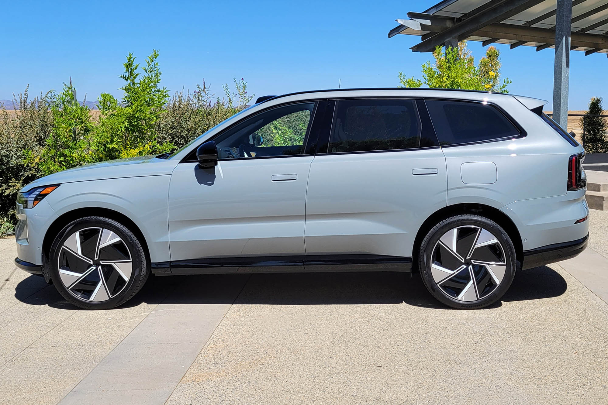 2025 Volvo EX90 Ultra in Vapor Grey parked on a driveway with shrubs and blue sky in the background.