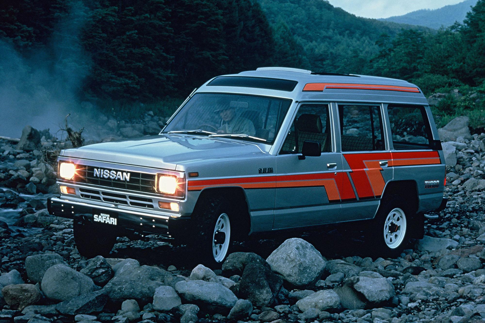 1983 Nissan Safari JDM vehicle parked on rocks with mountains in the background.