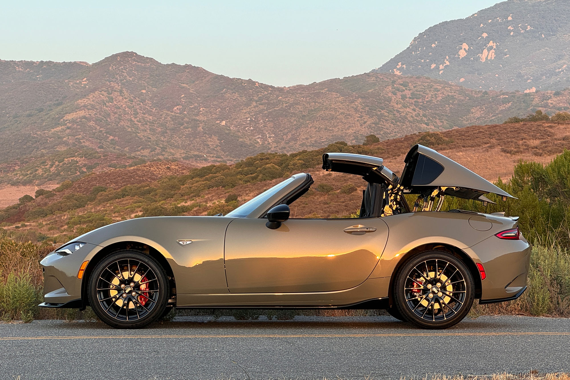 Side view of a 2024 Mazda MX-5 Miata RF Club in Zircon Sand, parked on a road at sunset with the top partially open and mountains behind it.