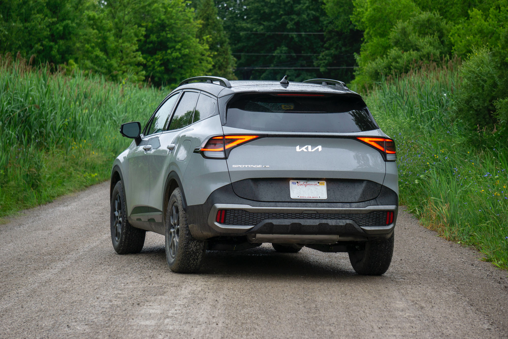 Rear-quarter of a Wolf Gray 2024 Kia Sportage X-Pro Prestige parked on a dirt road with trees in the background