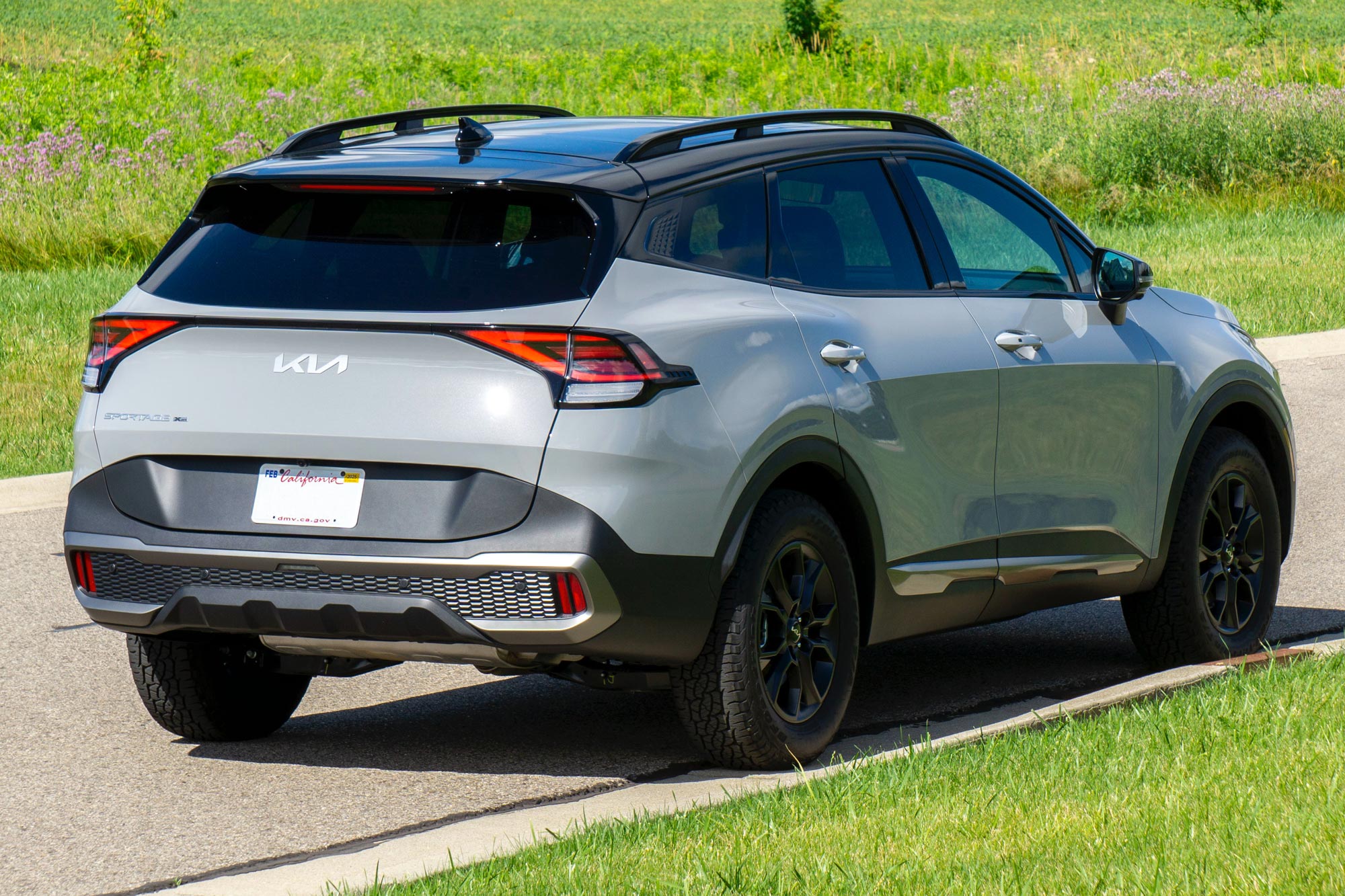 Rear-quarter of a Wolf Gray 2024 Kia Sportage X-Pro Prestige parked on a street with a field in the background