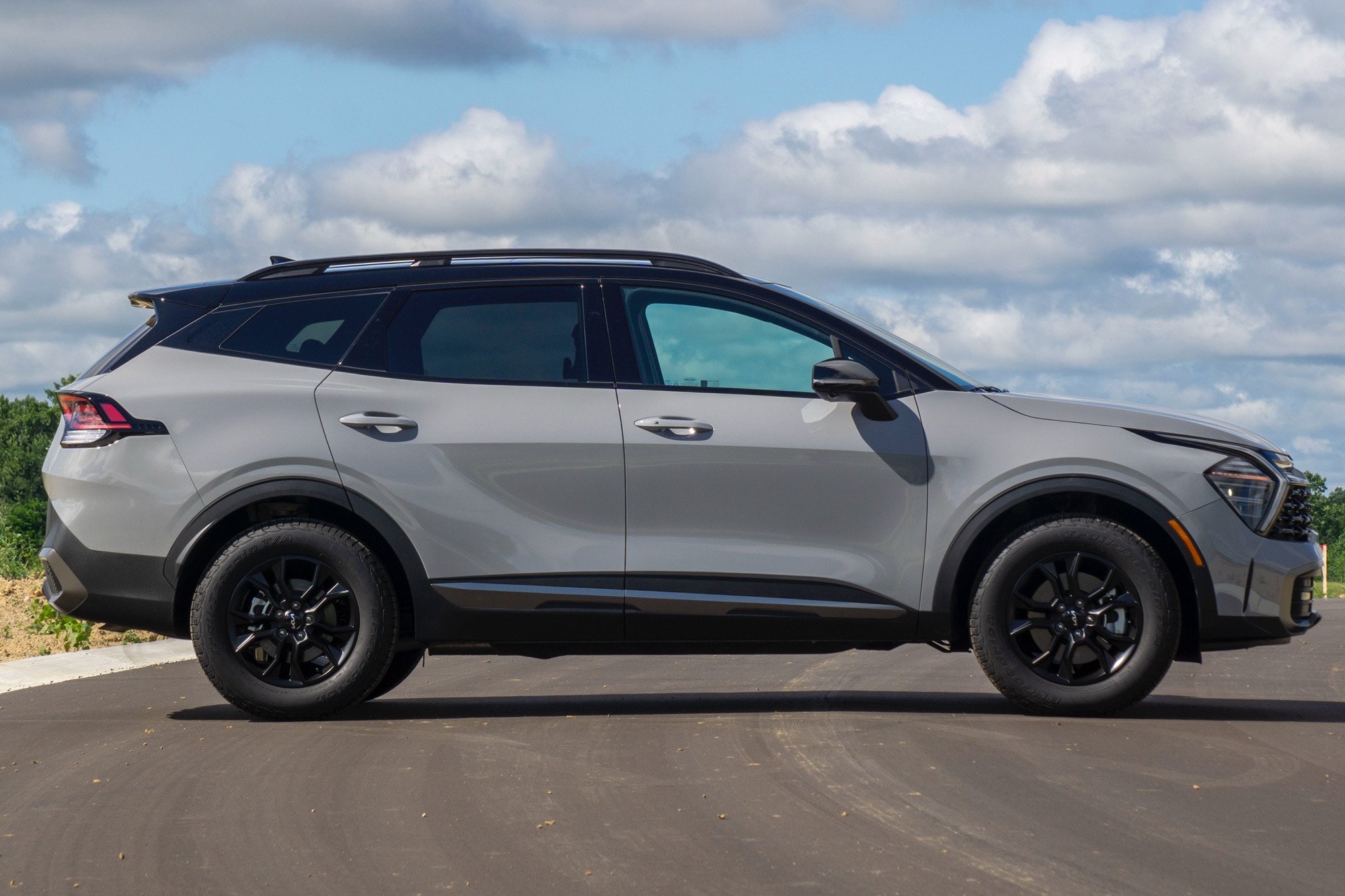 Side view of a Wolf Gray 2024 Kia Sportage X-Pro Prestige parked on a street with trees, blue sky, and white clouds in the background