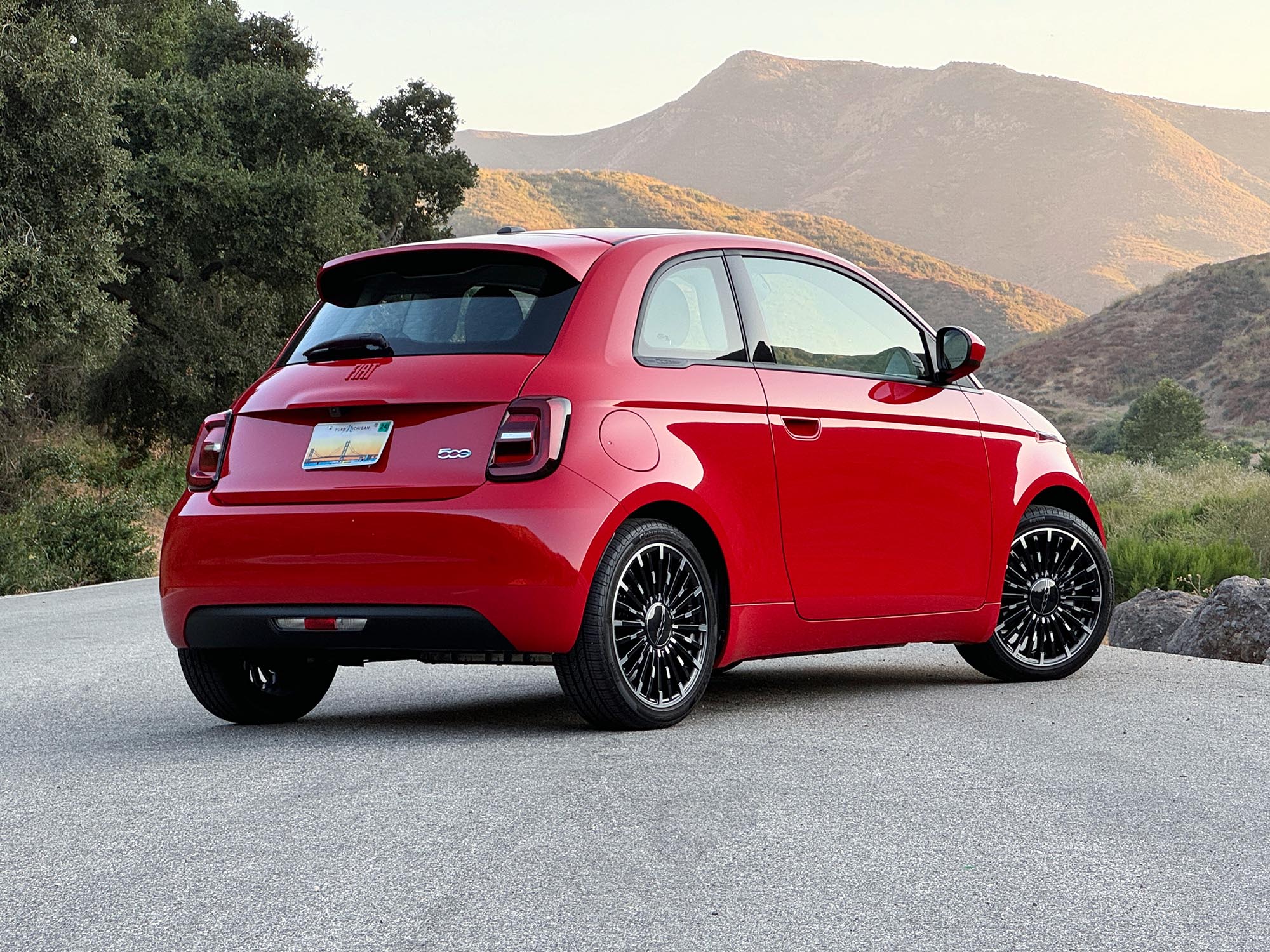 2024 Fiat 500e in red parked on a road at sunset with trees and mountains in the background, rear view