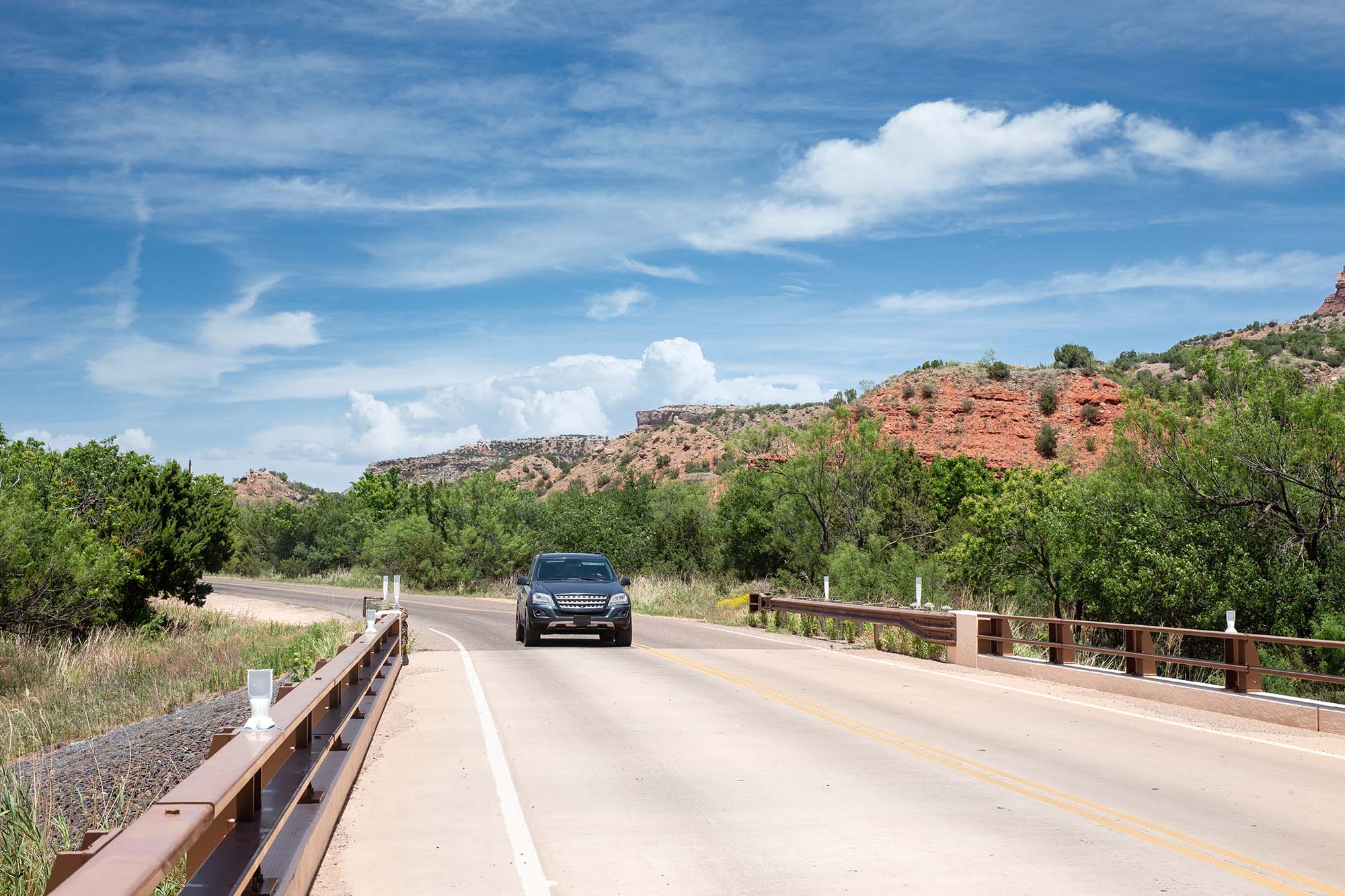 SUV drives down a hill on a paved road with red rock formations in the background.