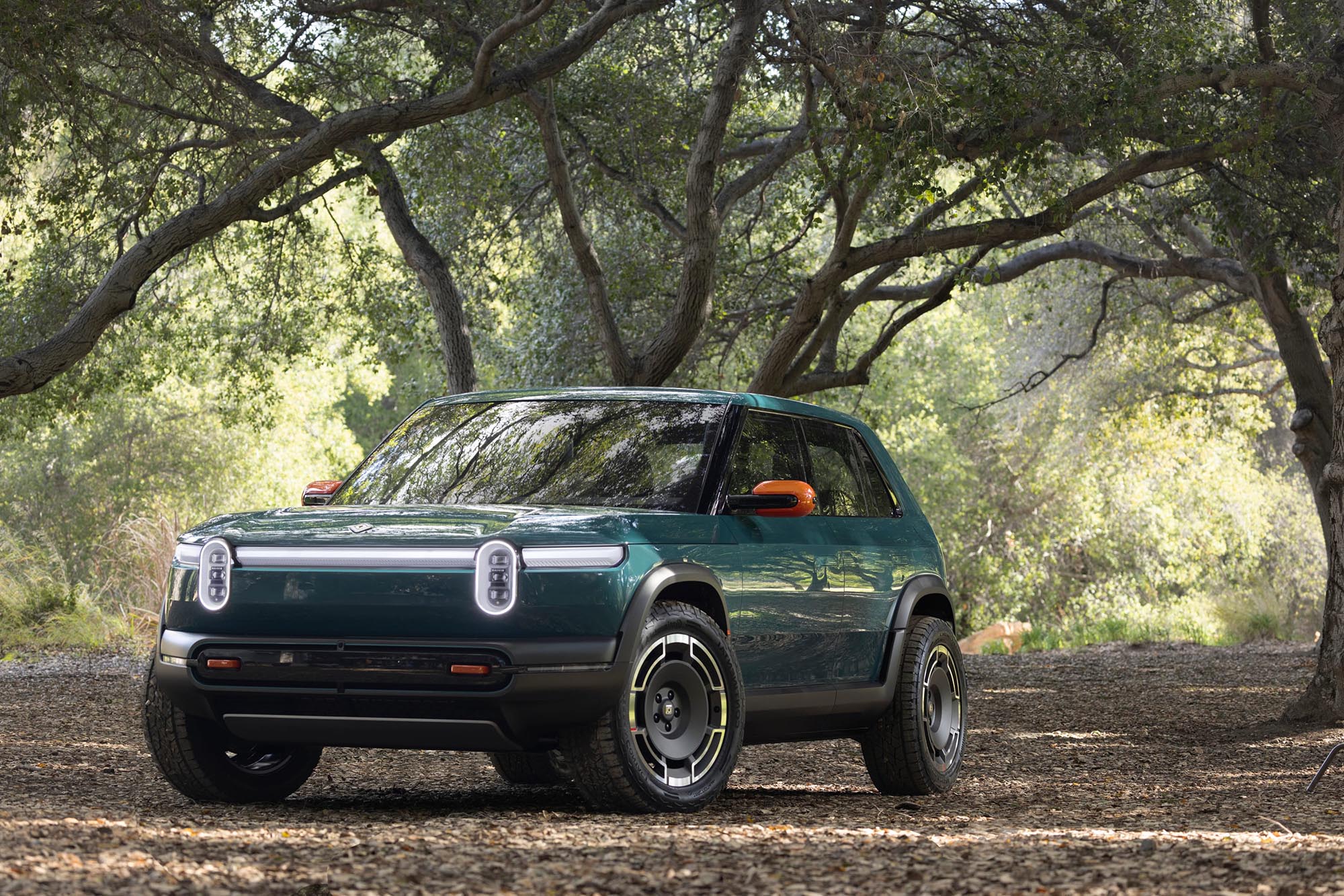 Green Rivian R3X parked on leaves beneath trees.