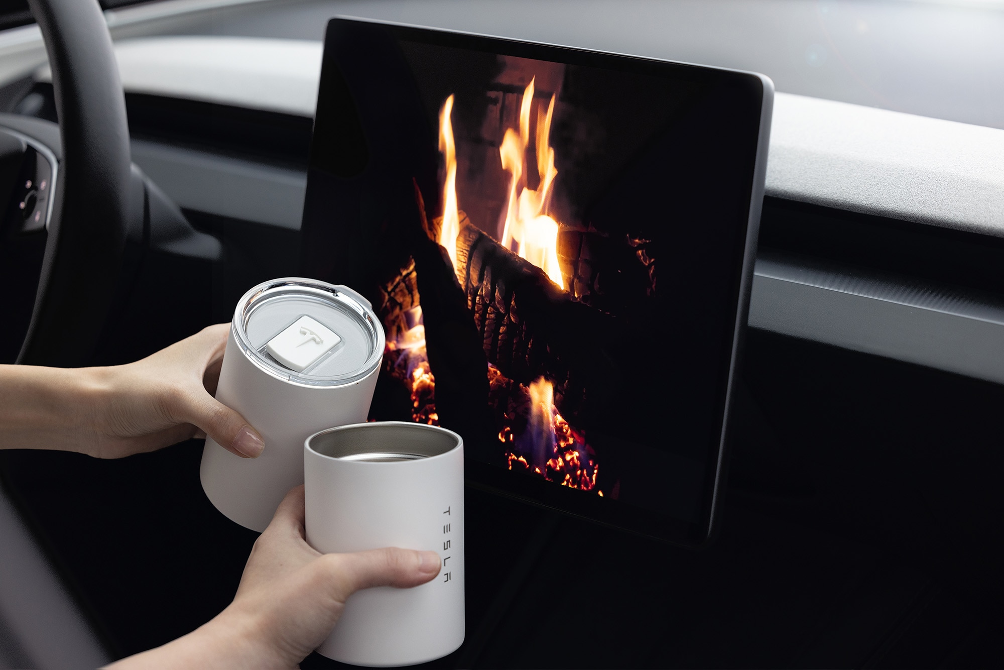  Interior of a Tesla Model 3 with a fireplace image on the infotainment screen and two hands holding white insulated tumblers.