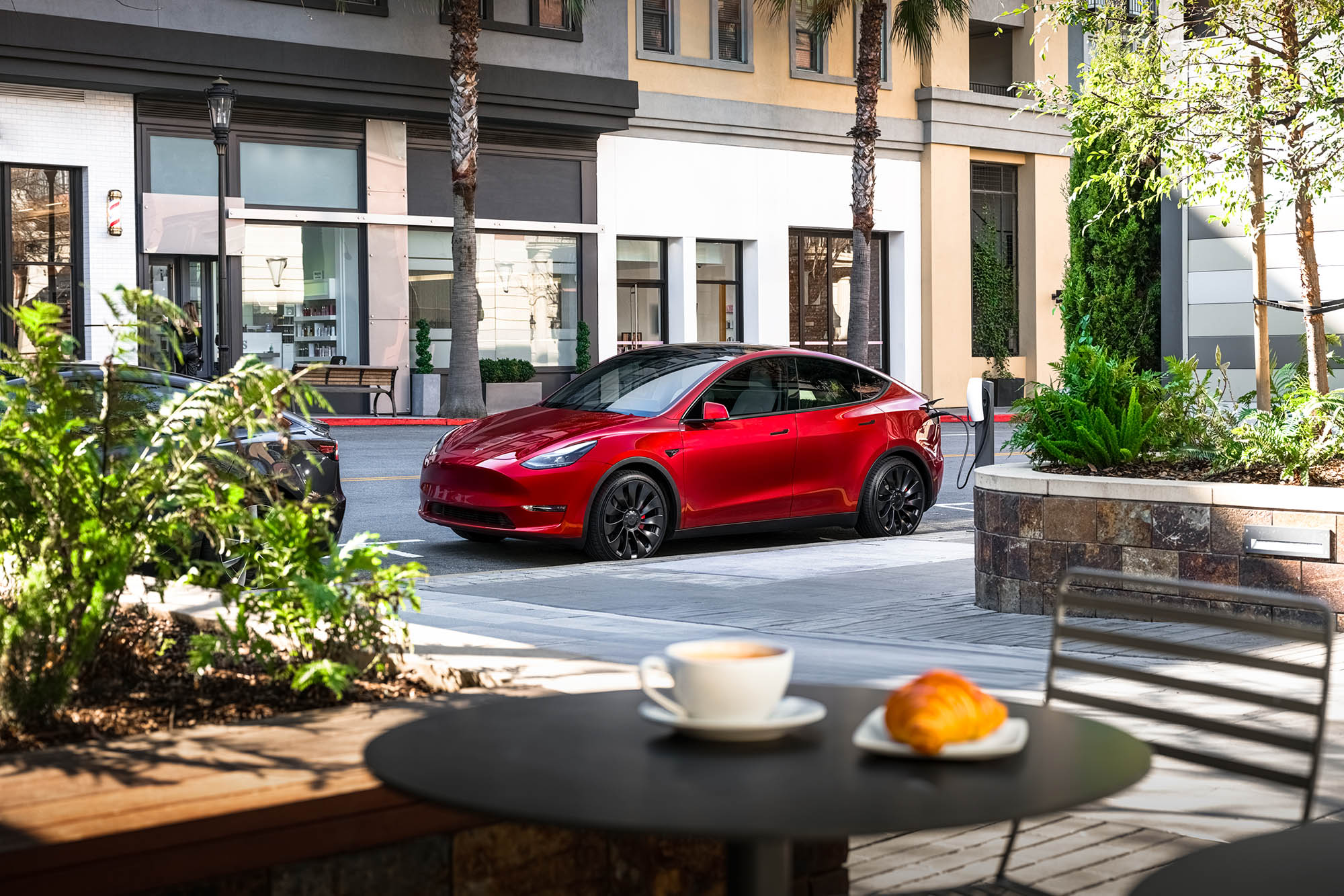 Red Tesla Model Y parked in front of a caf&eacute; with a table holding a coffee and croissant in the foreground.