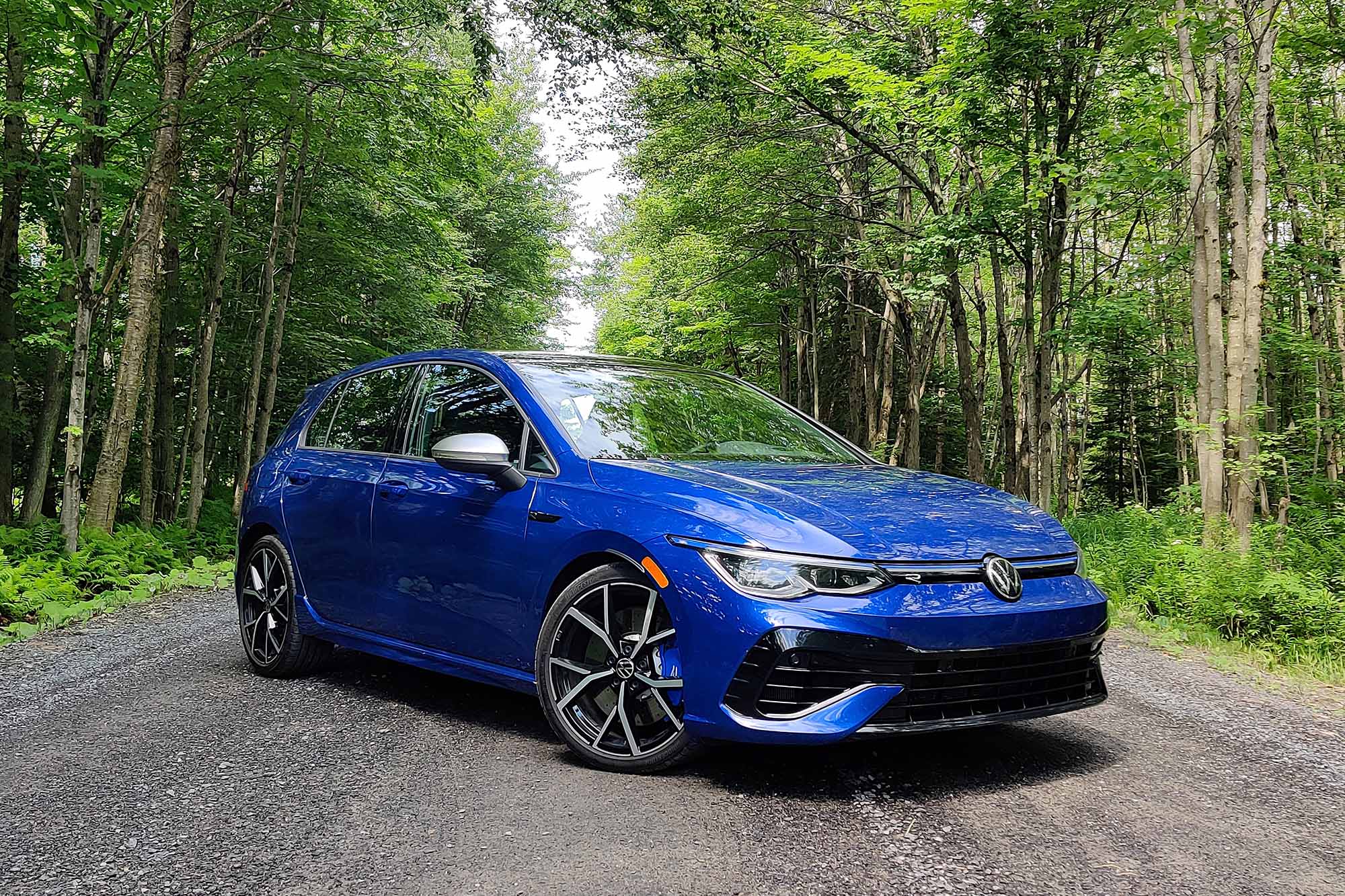 Front-quarter view of a blue 2024 Volkswagen Golf R parked on a gravel road in a forest.