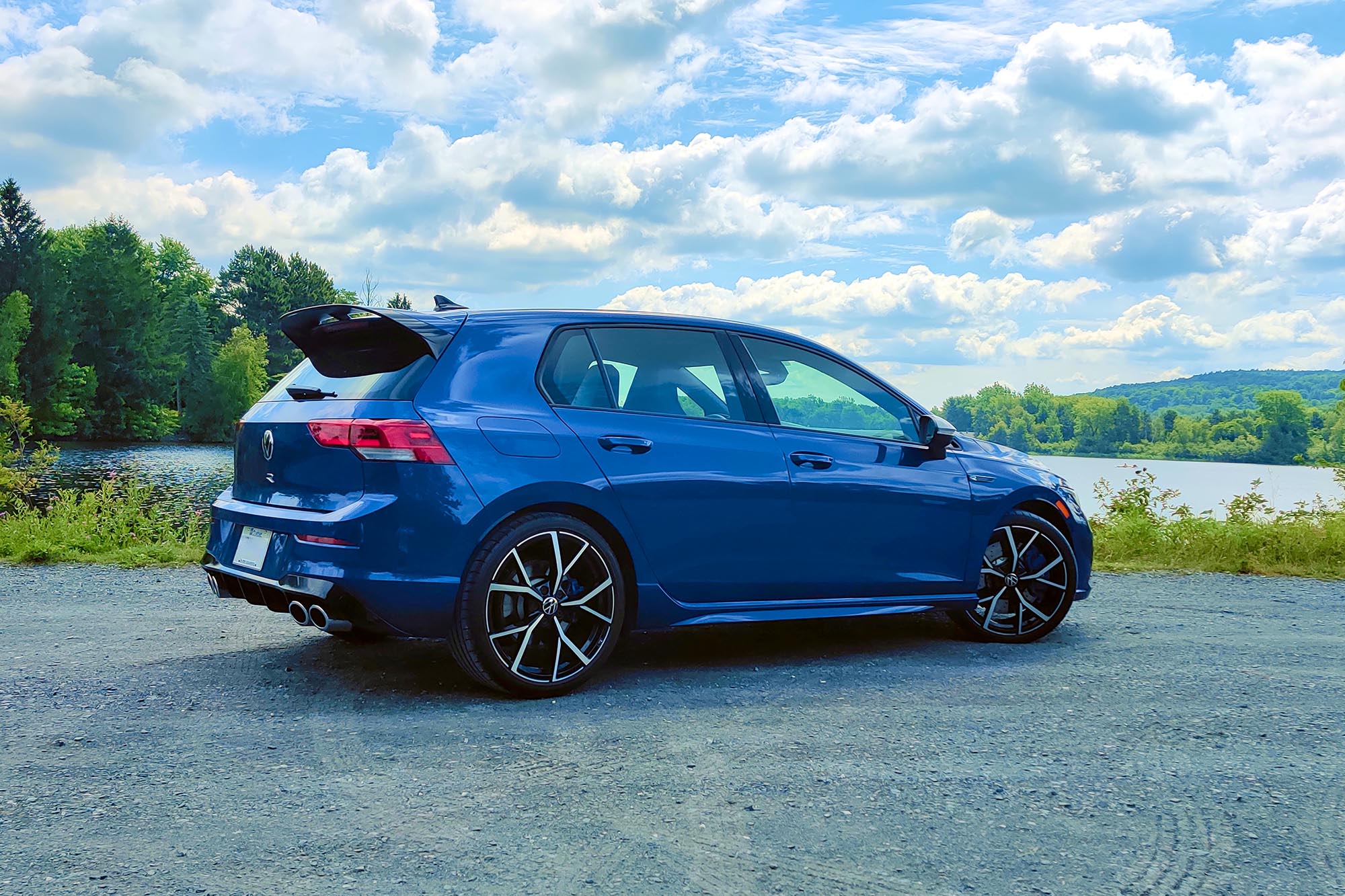 Rear-quarter view of a blue 2024 Volkswagen Golf R parked on a gravel surface with trees, hills, and a lake in the background.