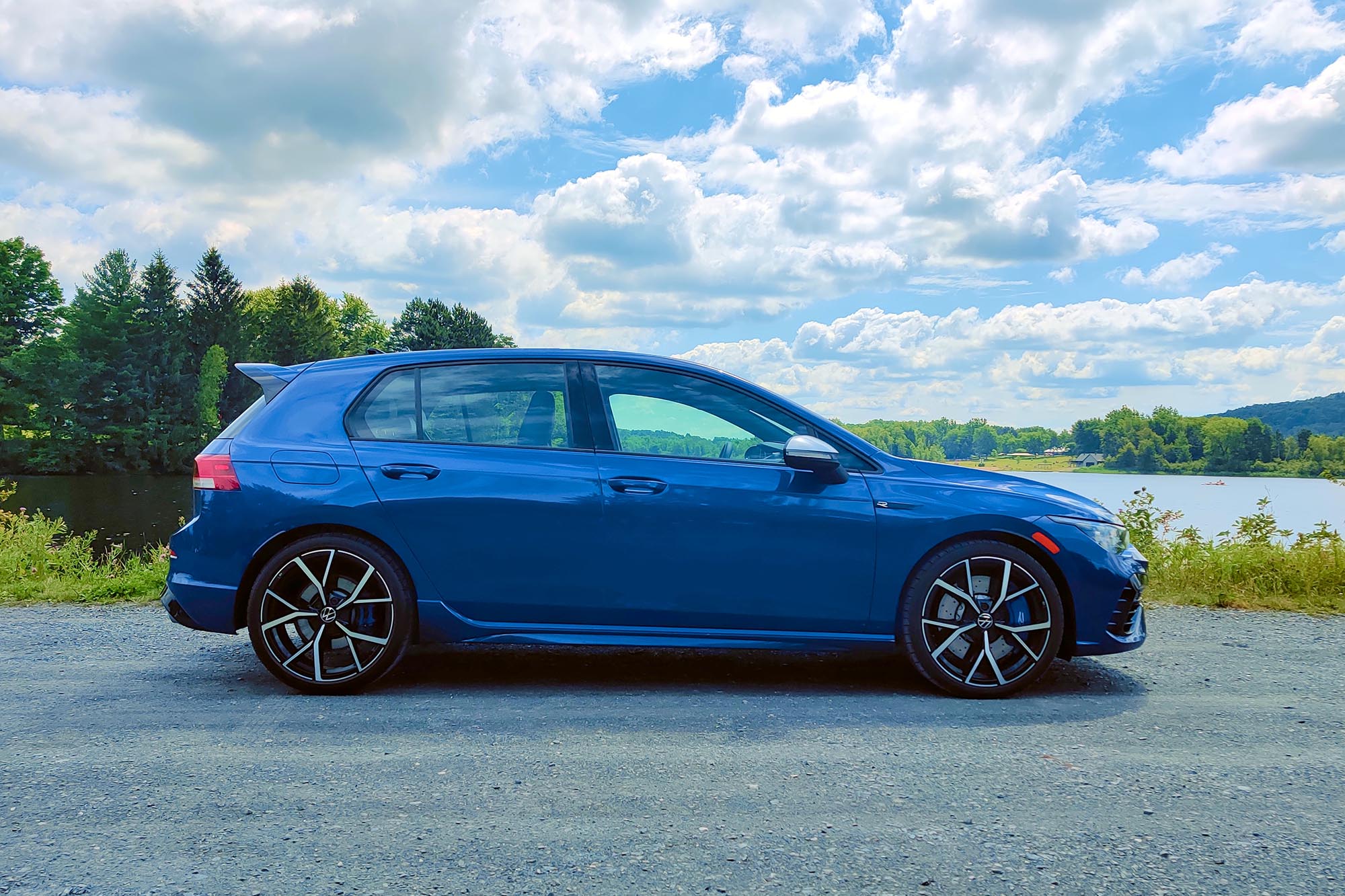 Side view of a blue 2024 Volkswagen Golf R parked on a gravel surface with trees and a lake in the background.