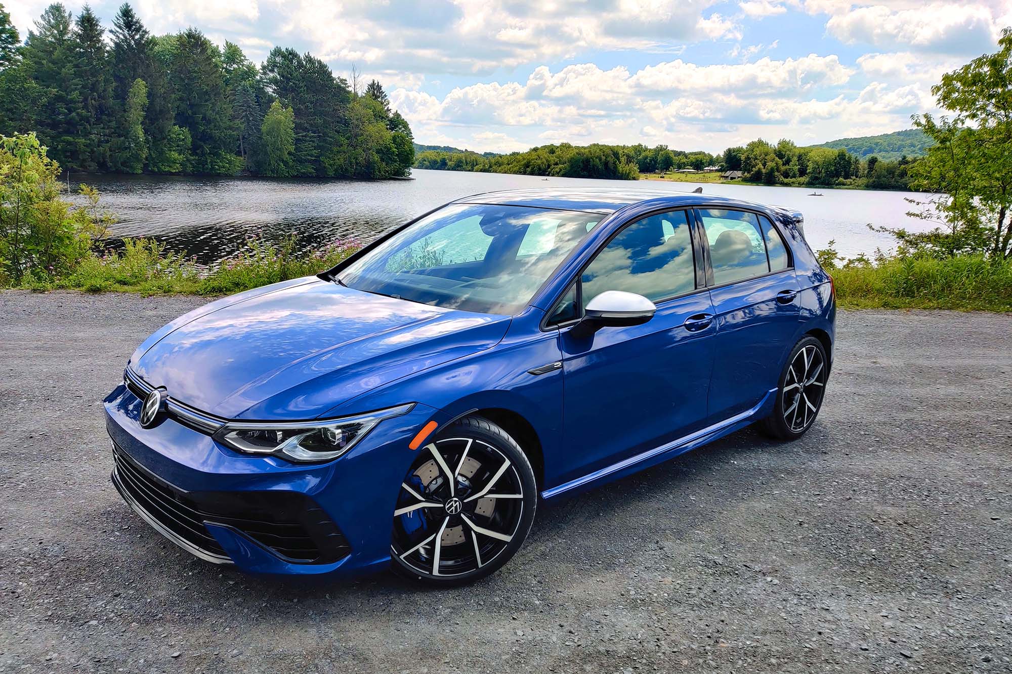 Front-quarter view of a blue 2024 Volkswagen Golf R parked on a gravel surface with trees, hills, and a lake in the background.