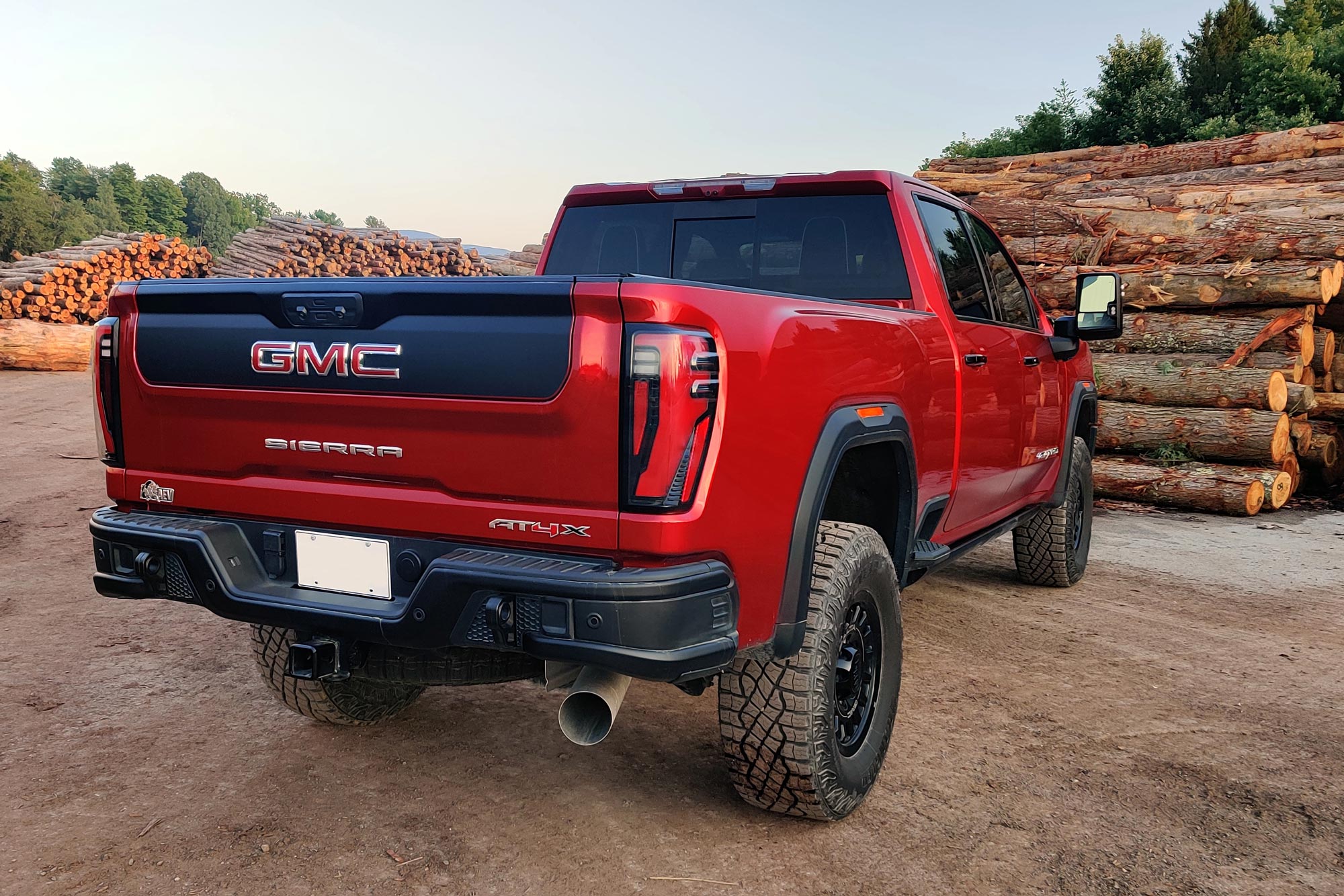 2024 GMC Sierra 2500HD AT4X AEV in Volcanic Red parked at a logging site with timber stacked behind it.