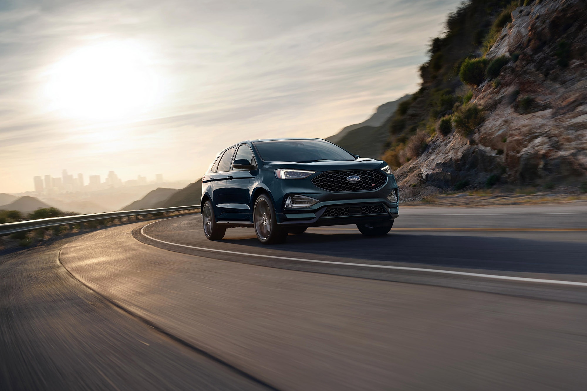 A blue-green Ford Edge driving in on a mountain road with the sun behind.