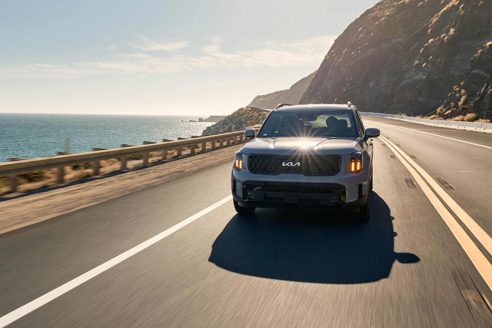 Gray Kia Telluride driving on a road with water to the side and mountains in the background.