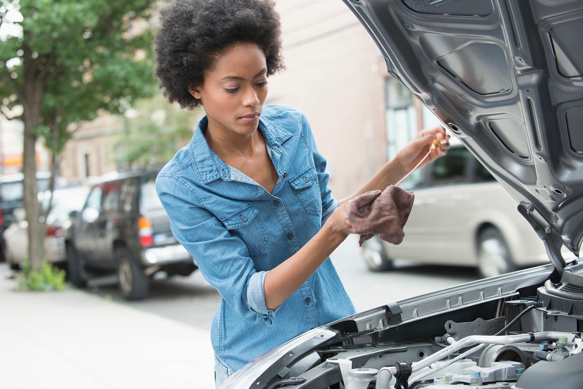 Person in a blue shirt looks at an oil dipstick next to a vehicle with its hood up.