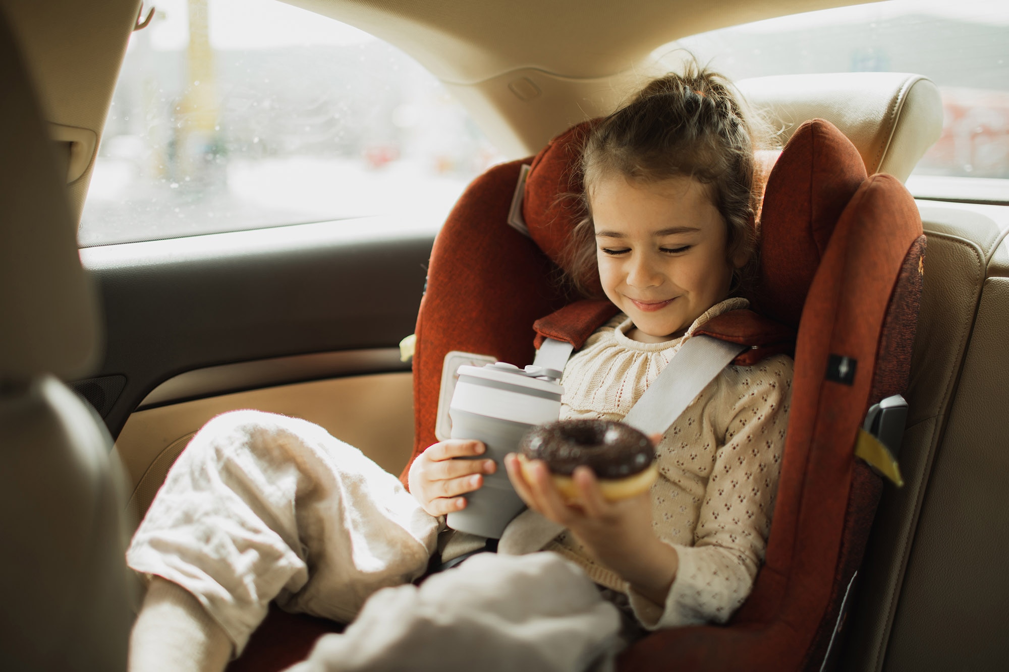 Child sits in forward-facing car seat holding a drink and a doughnut.