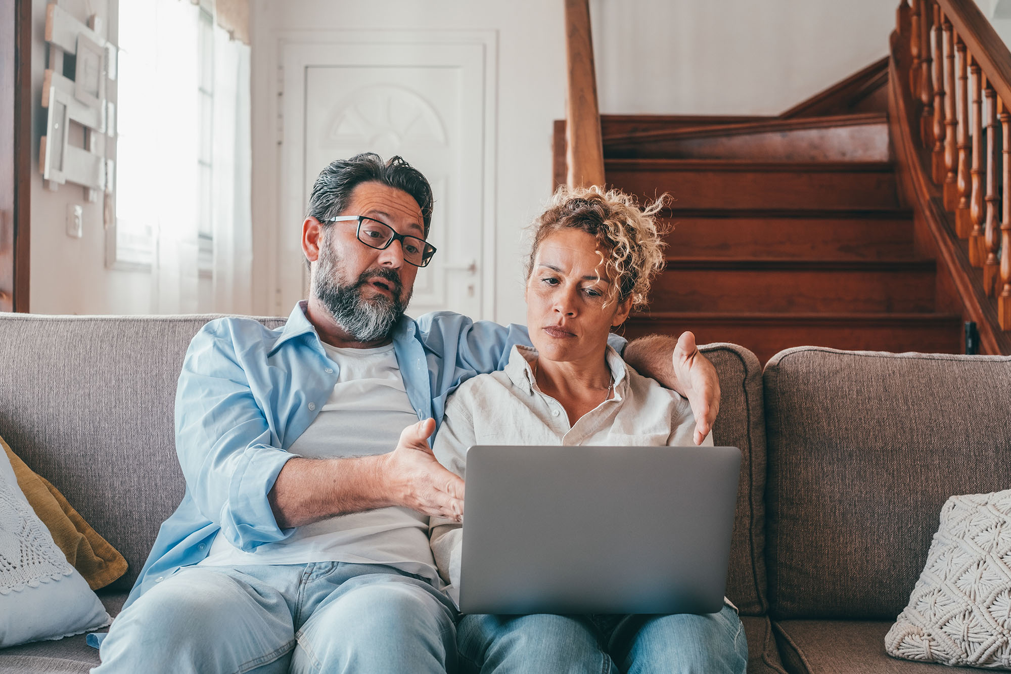 Two concerned-looking people sit on a couch with a laptop open in front of them.