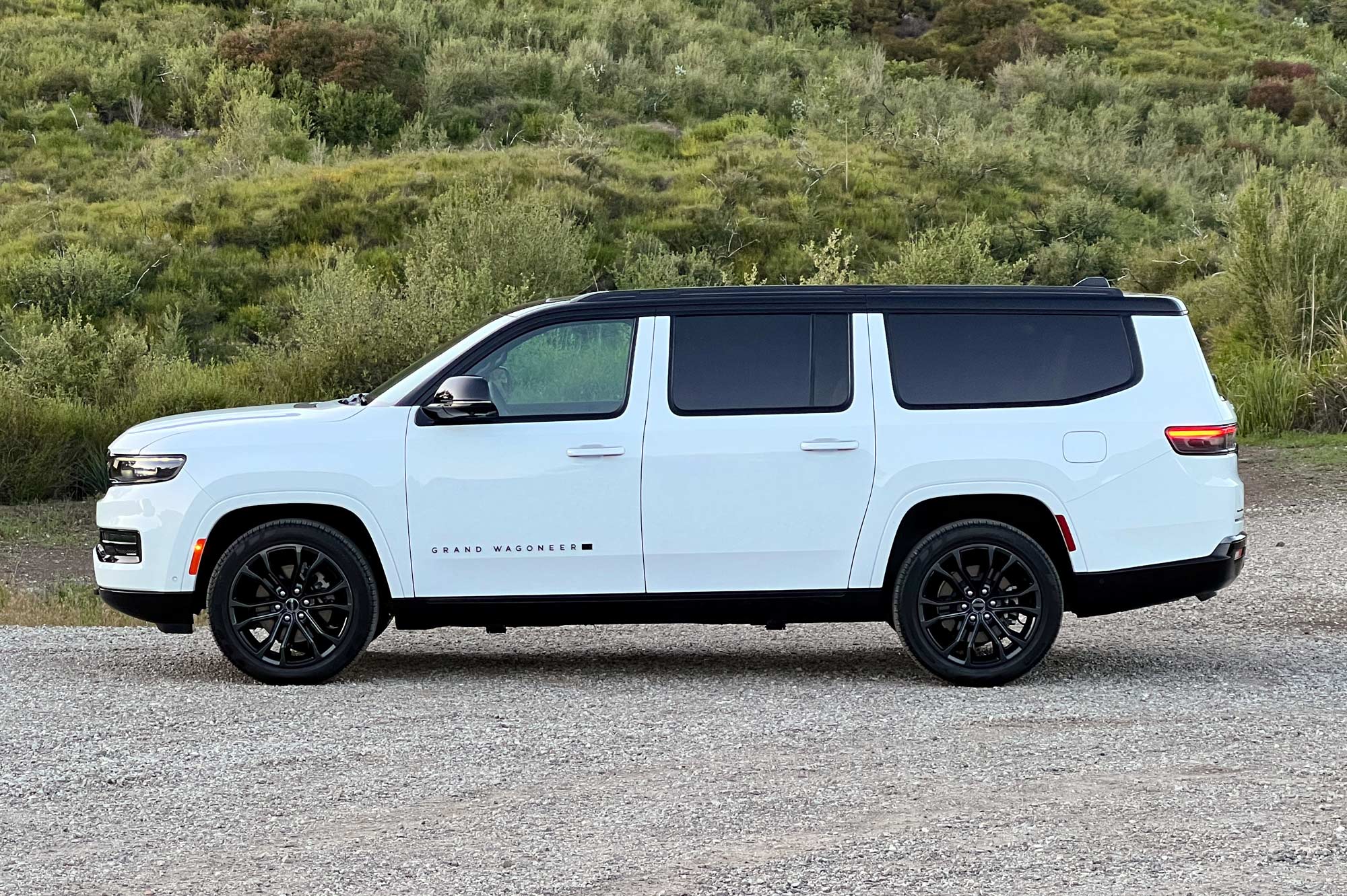 Side view of a white 2024 Jeep Grand Wagoneer L Series III Obsidian parked on gravel with green hills in the background