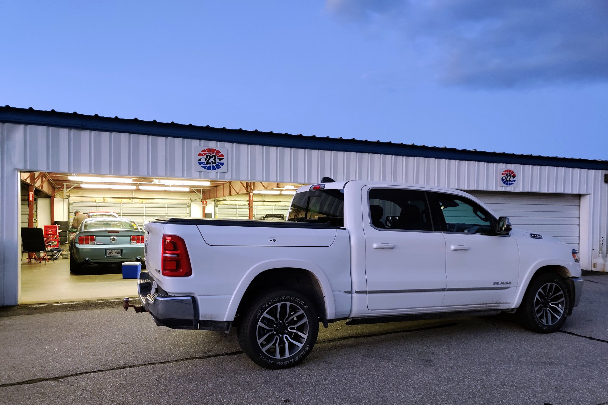 A blue Ford Mustang inside a garage with a white 2025 Ram 1500 outside