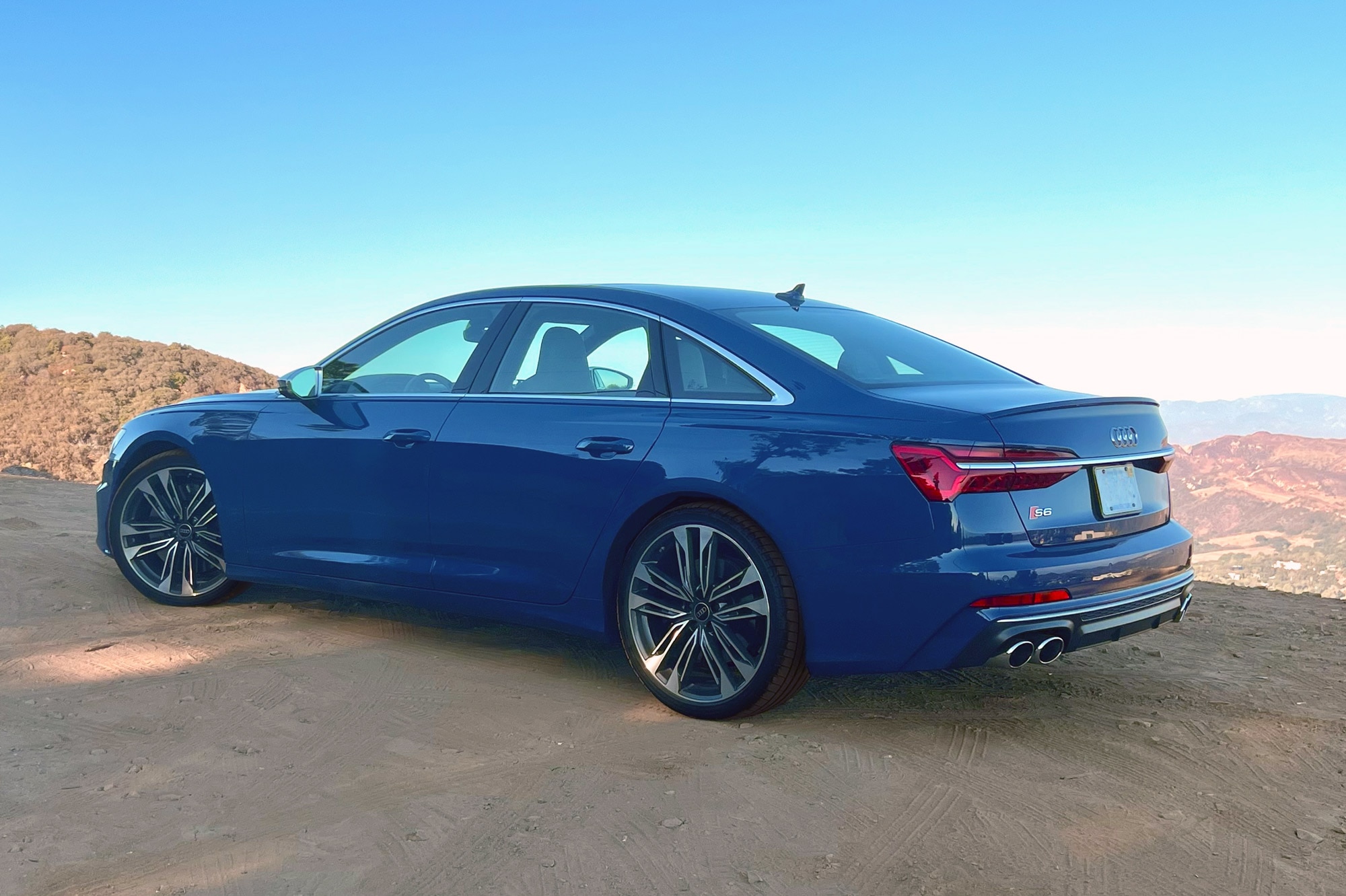 Rear quarter 2025 Audi S6 in Ascari Blue with rocks, brush, and a two-lane road in the background