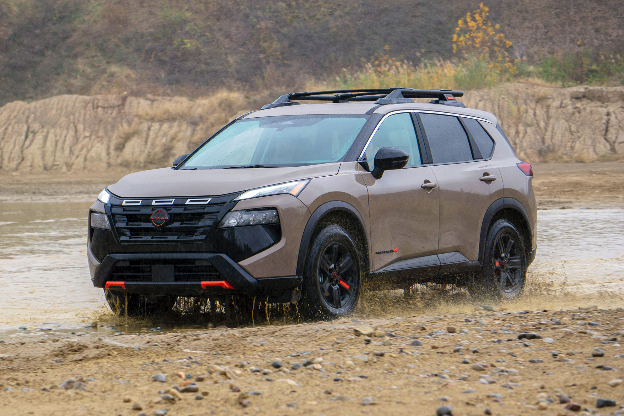 2025 Nissan Rogue Rock Creek in Baja Storm Metallic driving on the edge of a muddy stream.